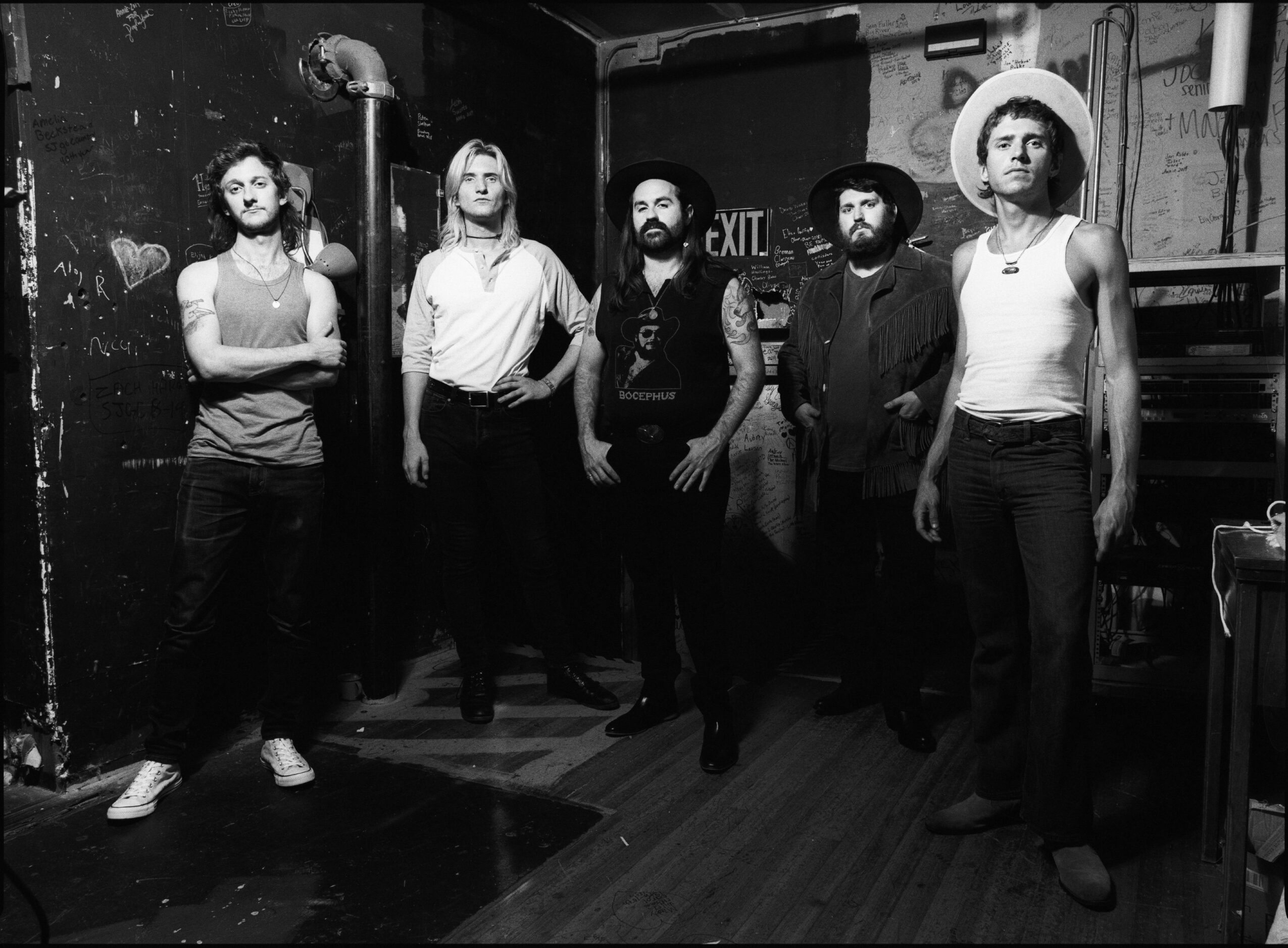Five people standing in a room with dark walls, some with arms crossed, one wearing a hat, in black and white.