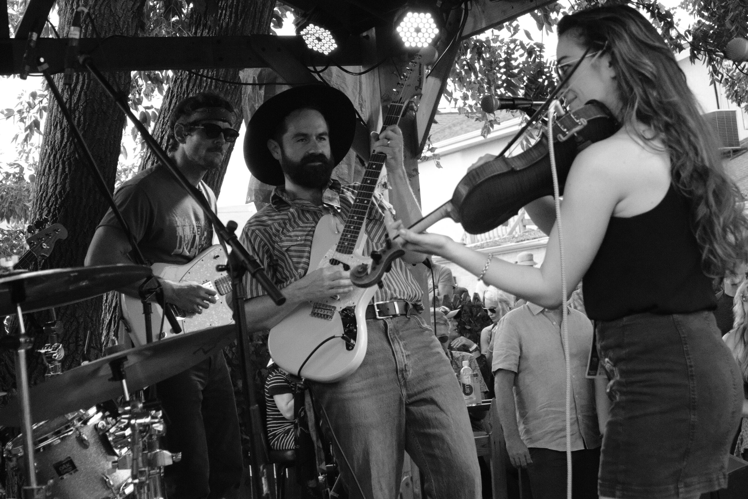 Three musicians perform outdoors, with a woman playing the violin and two men with guitars, under trees and string lights.
