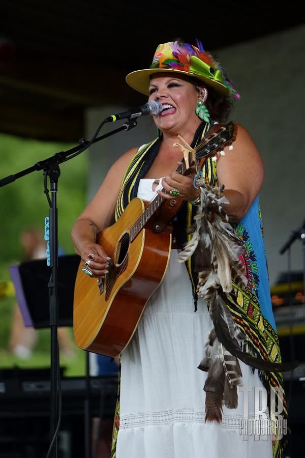 Woman wearing a colorful hat, singing and playing guitar on stage outdoors, smiling, with a microphone, in a white dress.