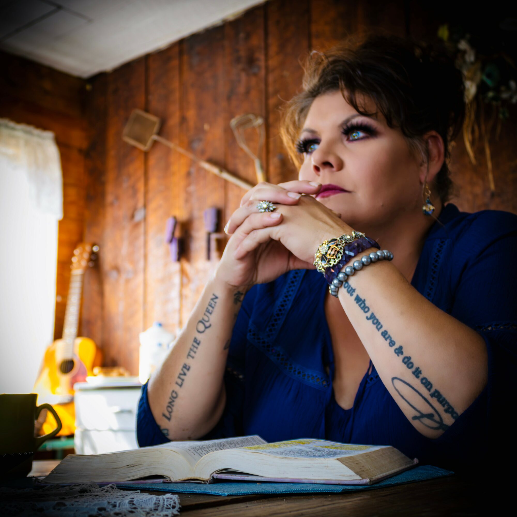 Woman with short hair and makeup resting her chin on her hands, sitting at a table with an open book, in a wooden room.