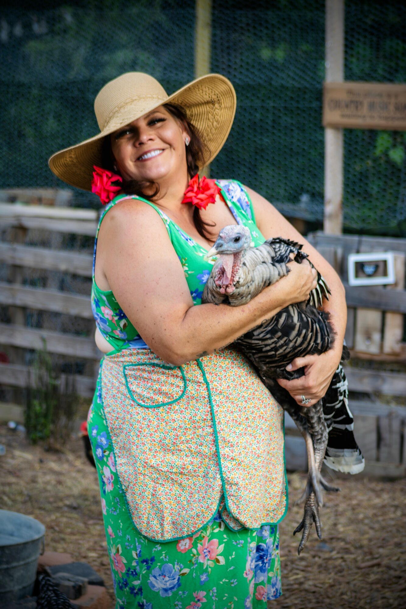 Woman in a wide-brimmed hat holding a turkey, smiling outdoors with a wooden fence in background.