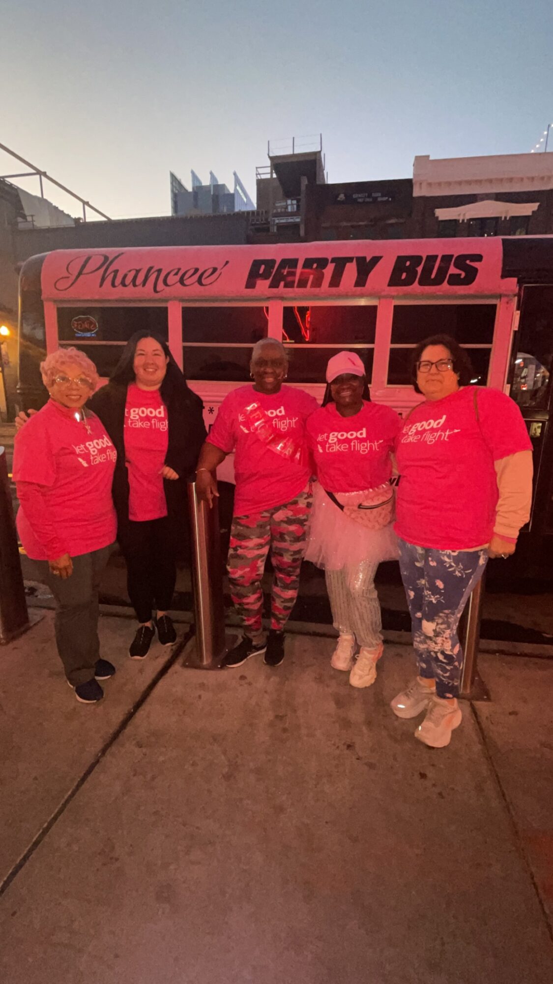 Five women in pink shirts stand in front of a pink party bus, smiling outdoors during daytime.
