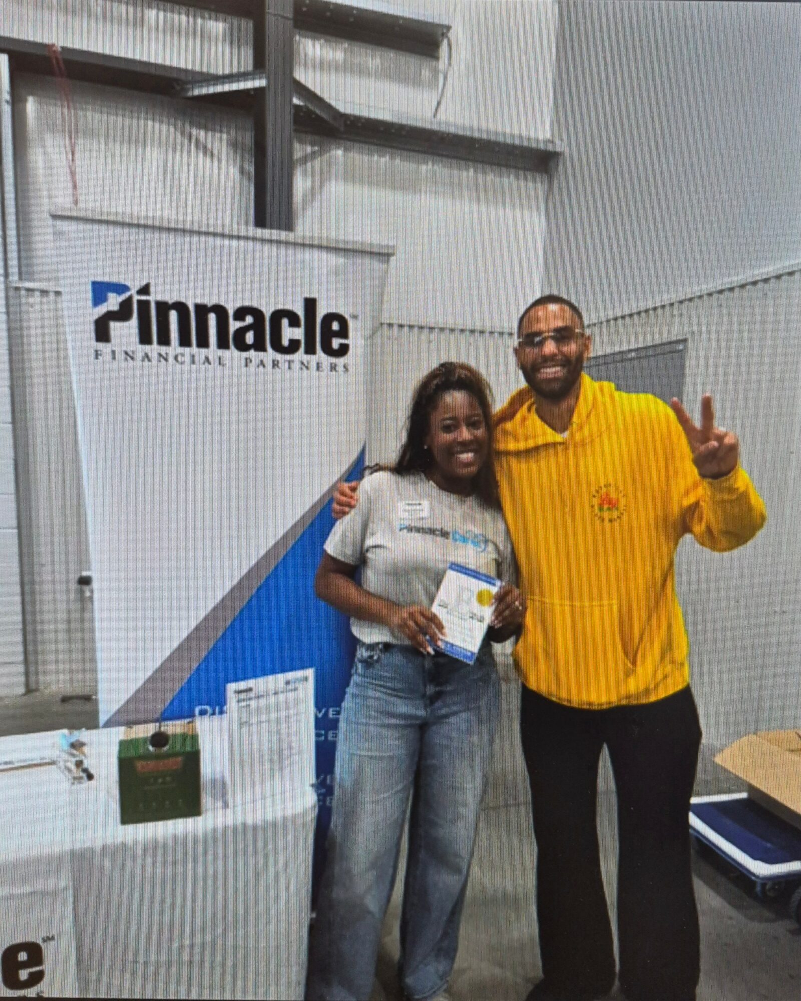 Two people standing indoors, one holding a certificate, smiling, with a Pinnacle Financial Partners banner in the background.