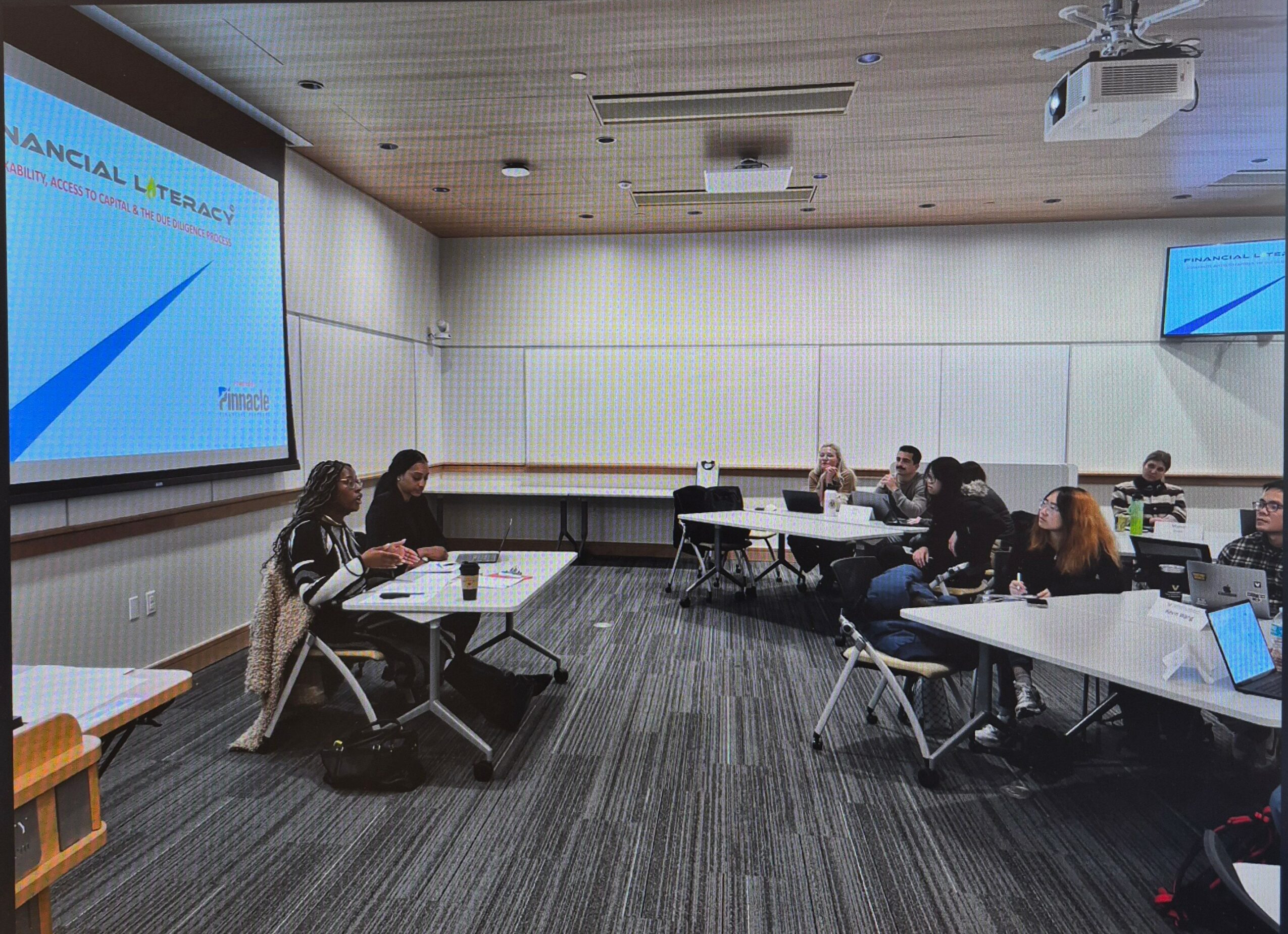 People seated at tables in a conference room with large screens on the walls, some using laptops.