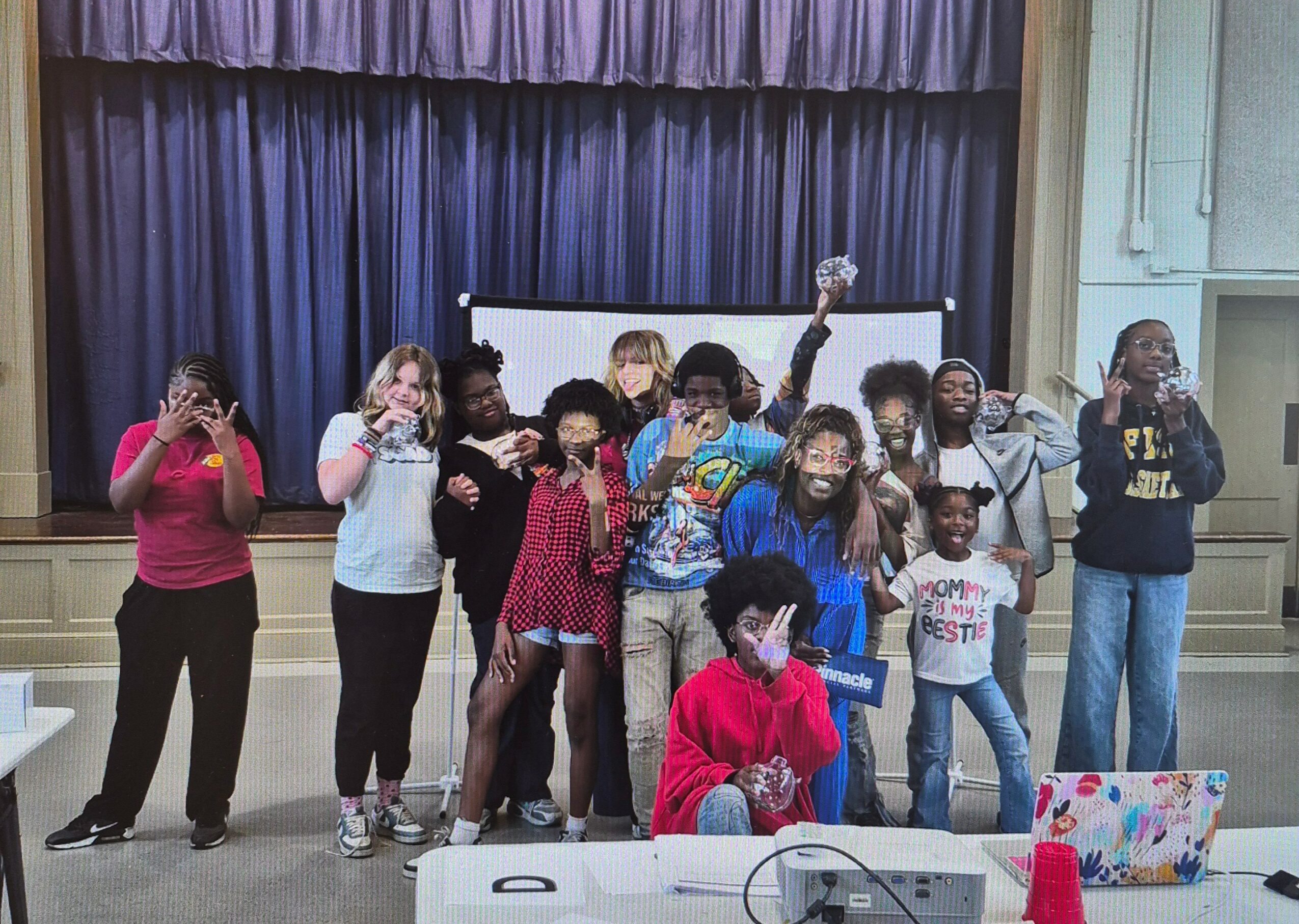 Group of children and teenagers standing on stage in front of a blue curtain, some holding phones, smiling and posing.