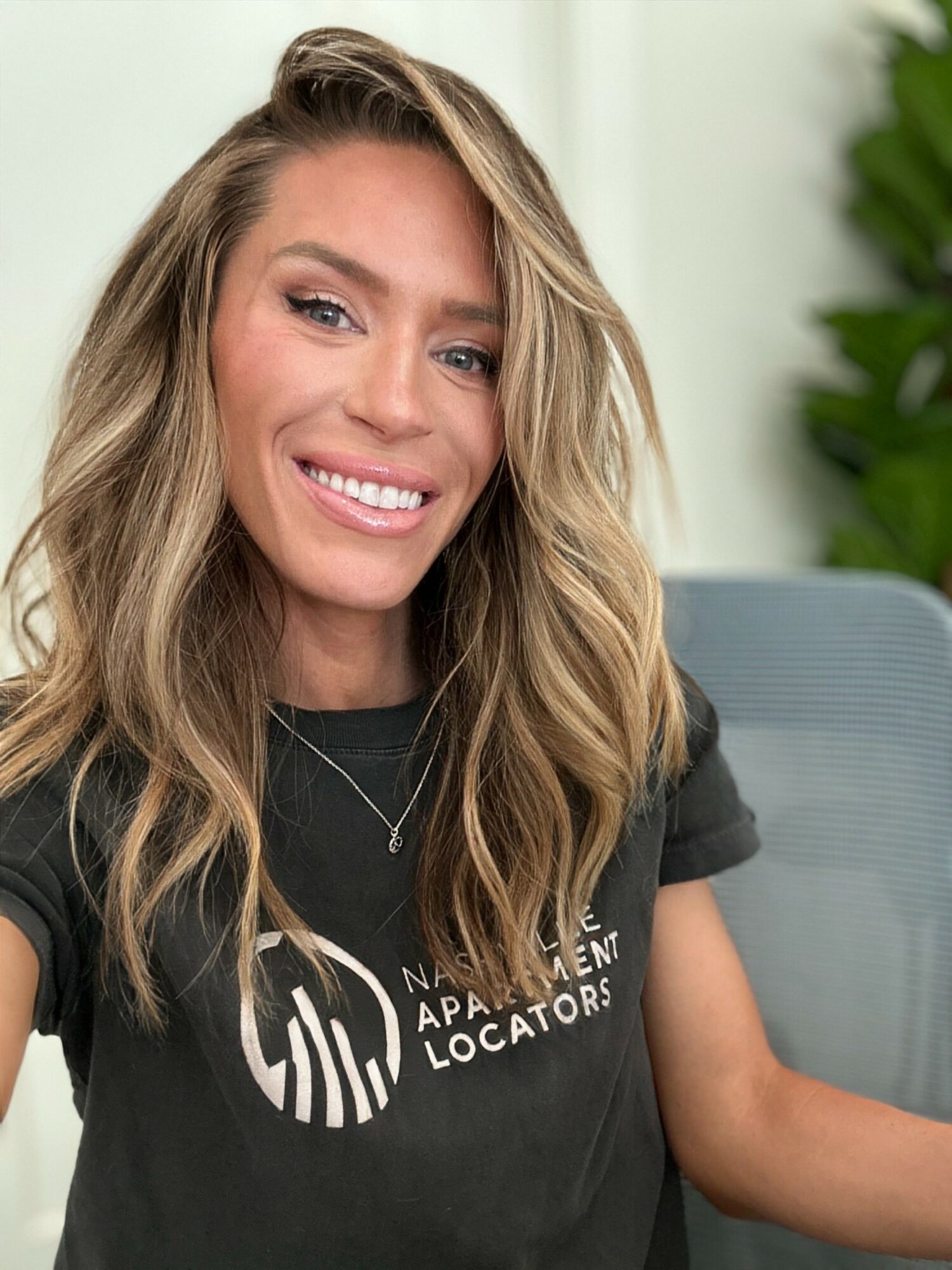 Smiling woman with long wavy hair wearing a black T-shirt with white text, sitting indoors near a plant.