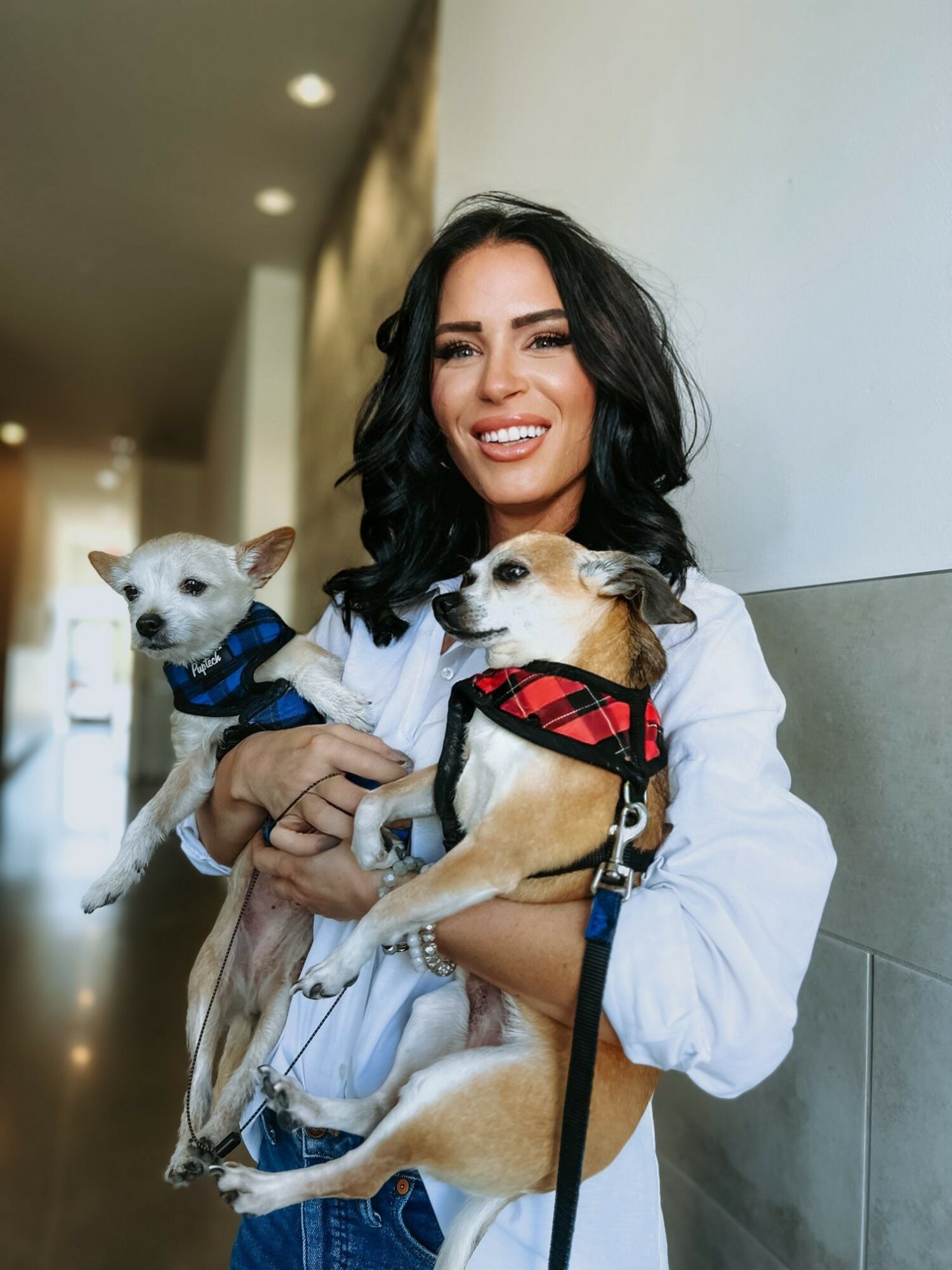 Woman with dark wavy hair smiling, holding two small dogs, one in each arm, indoors.