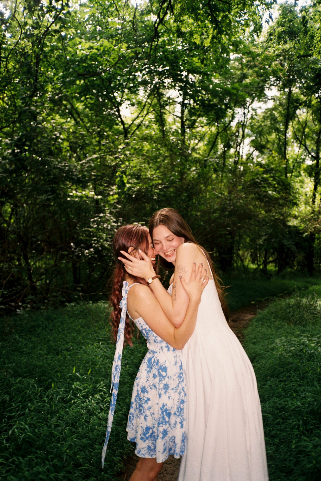 Two women hugging outdoors with trees and greenery in the background, one in a white dress and the other in a blue floral dress.