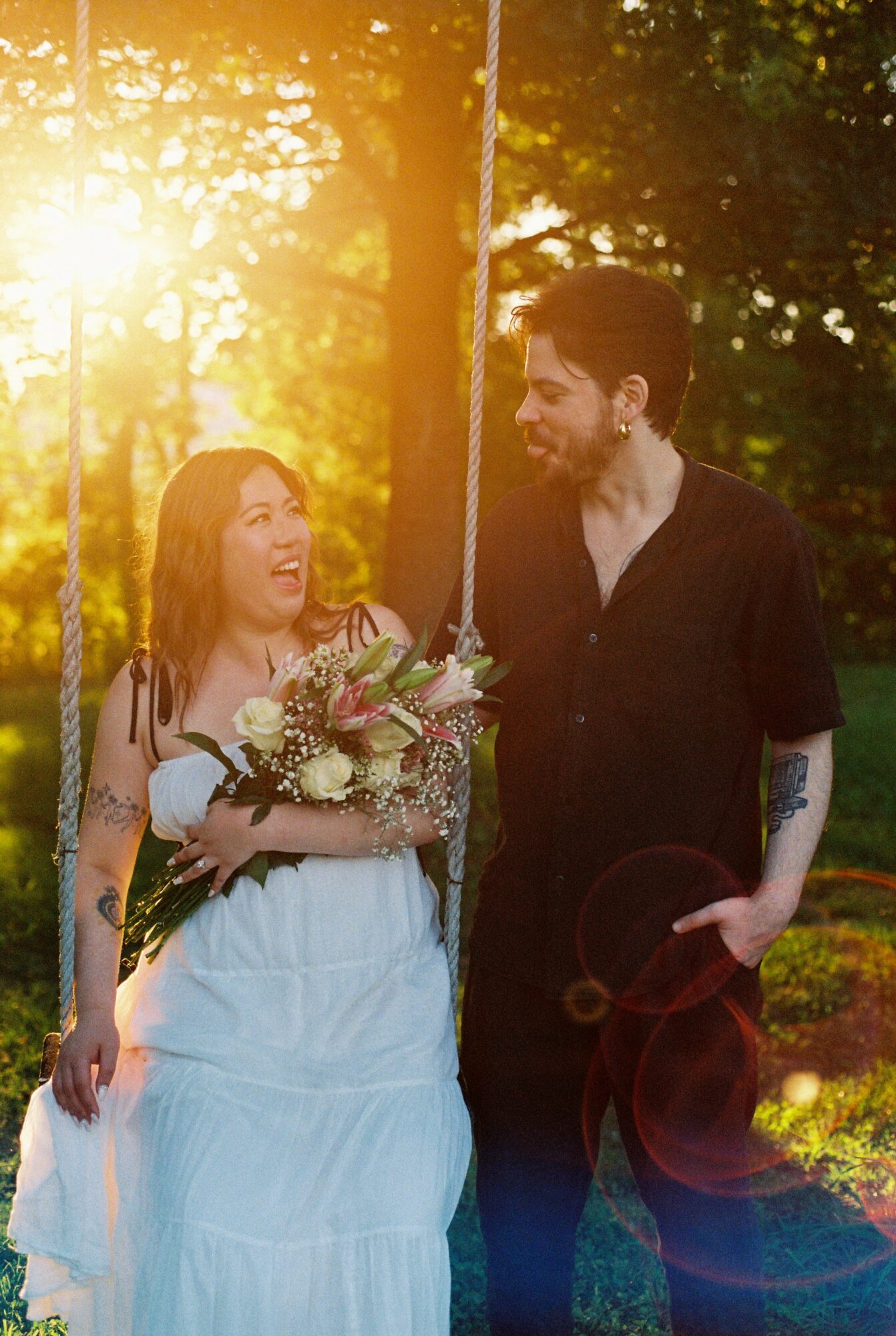A smiling woman in a white dress holding a bouquet, standing next to a man in a black shirt outdoors during sunset.