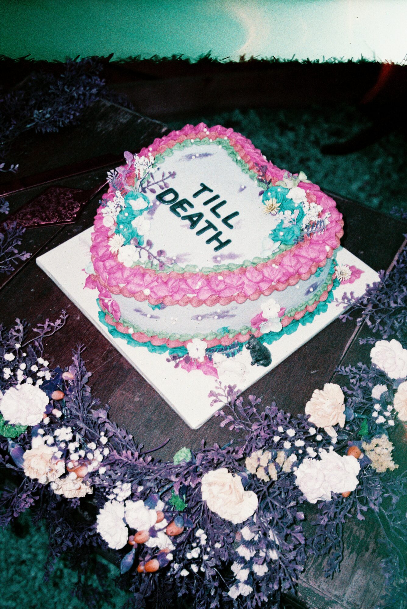 Heart-shaped cake with pink, blue, and white decorations, inscribed with 'TILL DEATH' on top, surrounded by flowers.