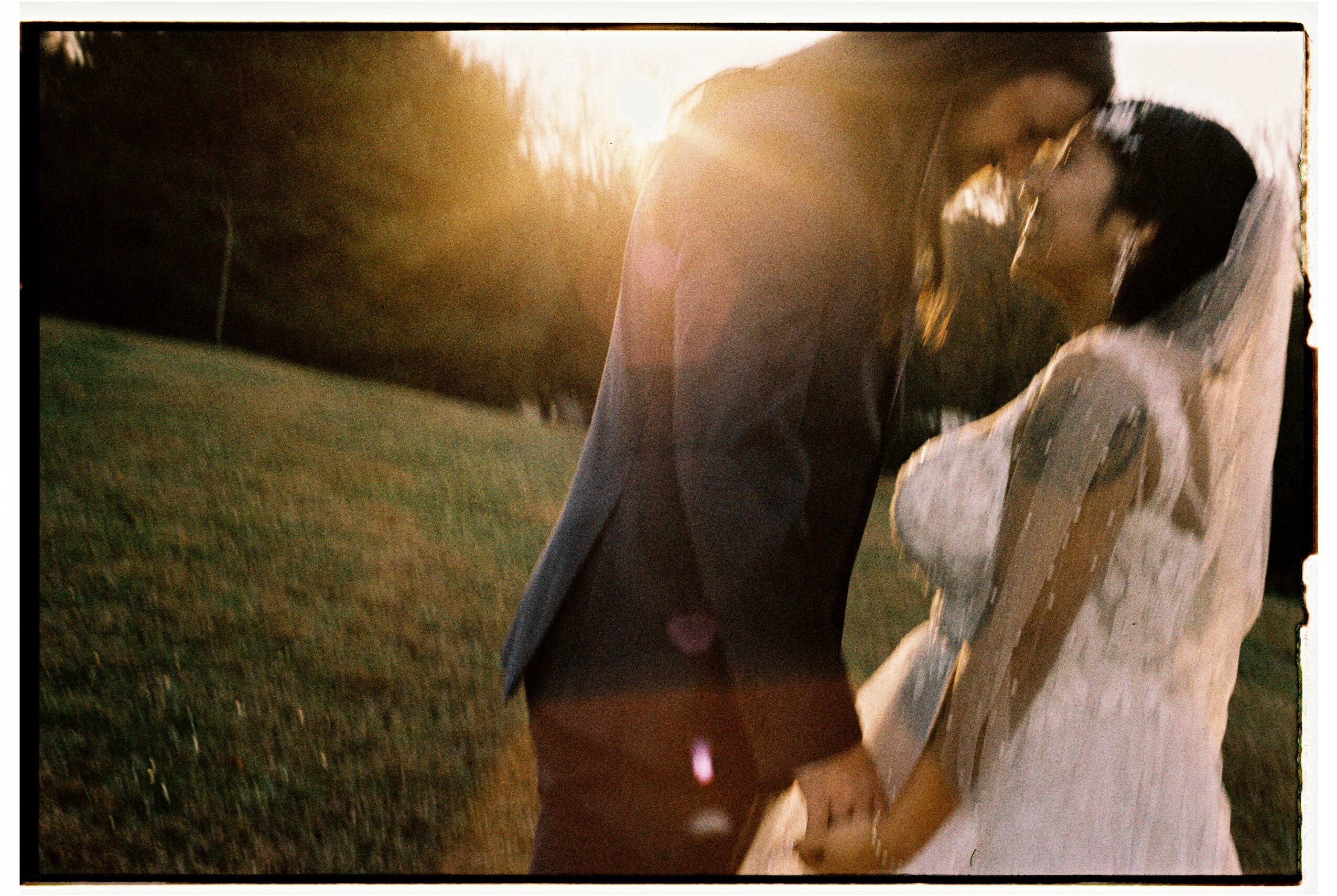 Couple holding hands outdoors at sunset, with trees in the background, embracing and about to kiss, wearing wedding attire.