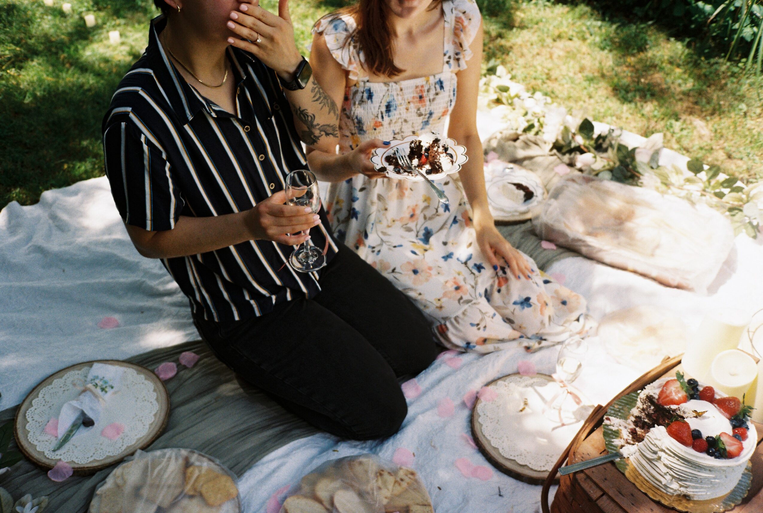 Two women having a picnic on a blanket outdoors with a cake and desserts, one holding a glass of wine, surrounded by greenery.
