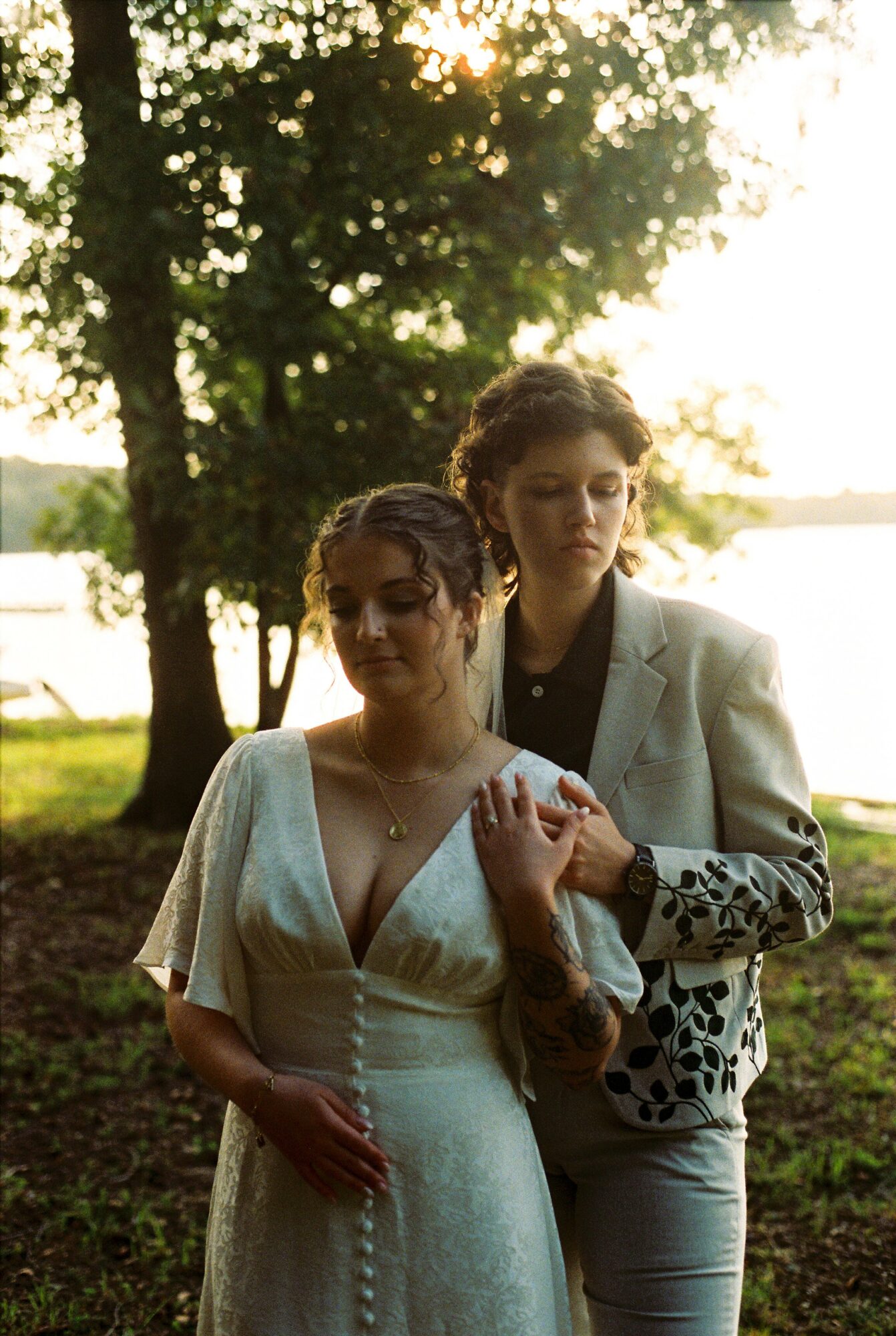Two women standing outdoors near a lake with trees, one in a white dress and the other in a light suit, during sunset.