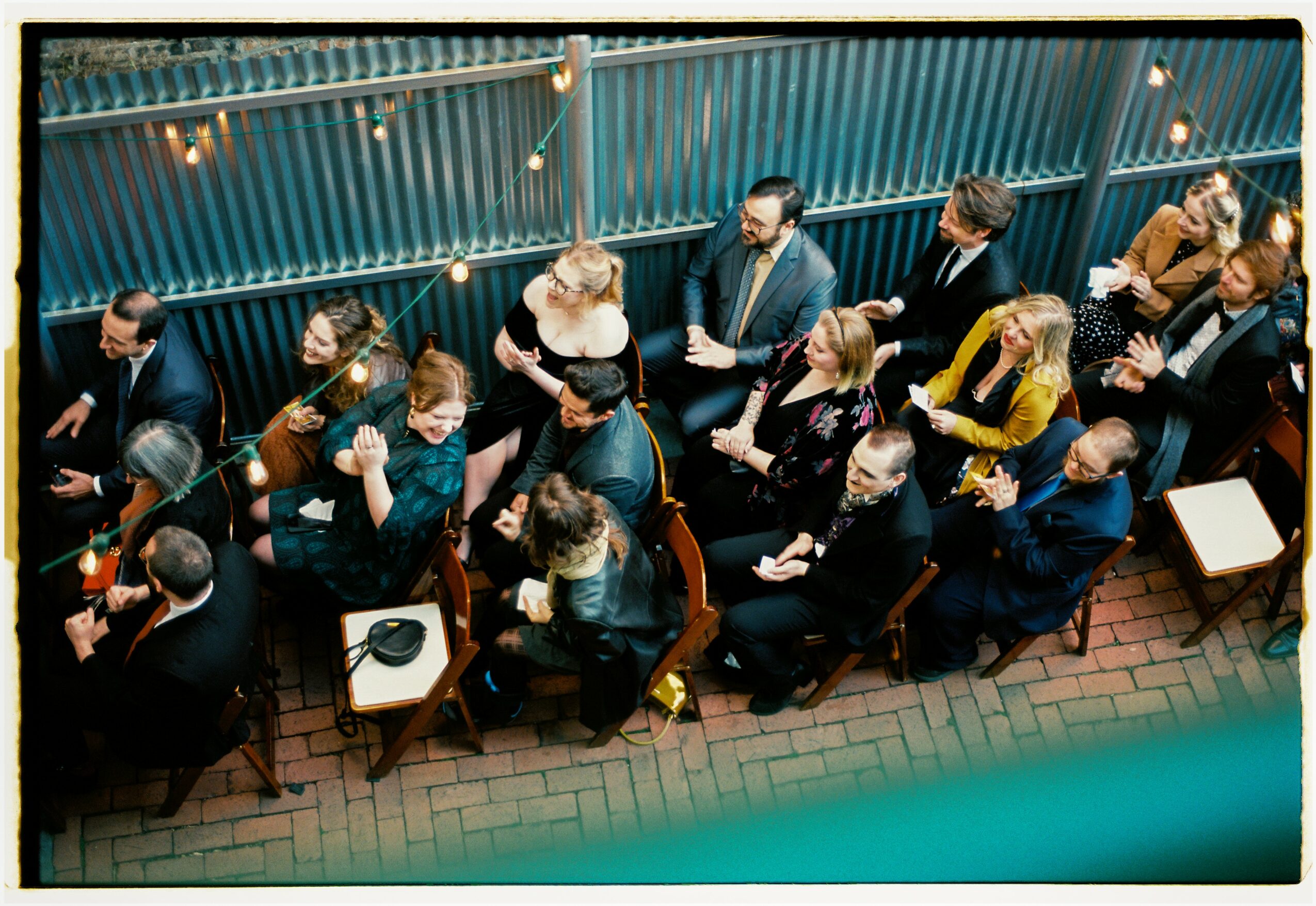 People sitting at tables outdoors, viewed from above, with string lights overhead and a blue fence background.