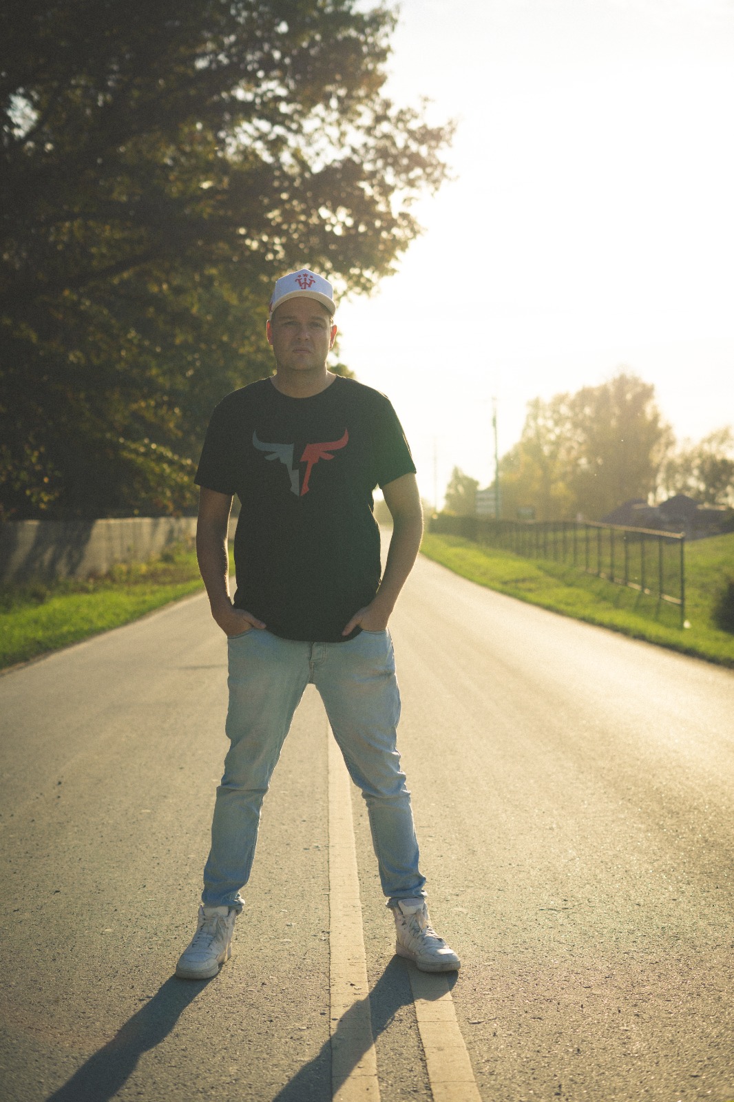 Person standing in the middle of a road with trees and a fence in the background, sunlight behind them, hands in pockets.