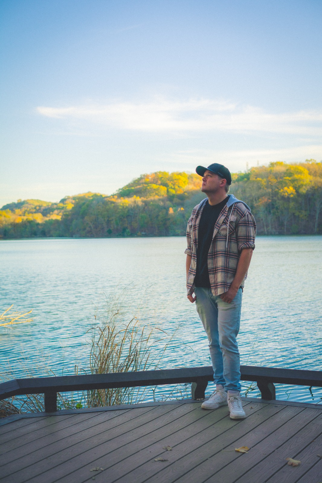 Young man standing on a wooden dock by a lake with trees and hills in the background, under a blue sky with clouds.