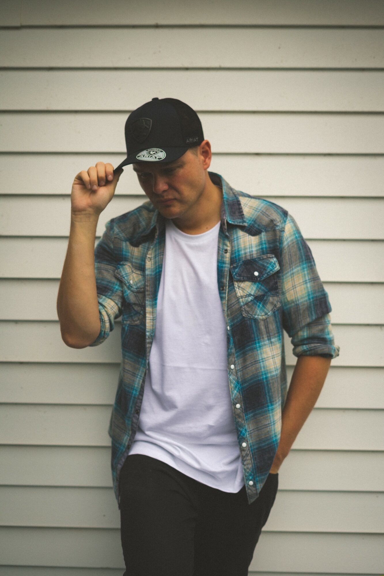 Young man in plaid shirt and baseball cap adjusting cap, standing against a white horizontal siding wall.