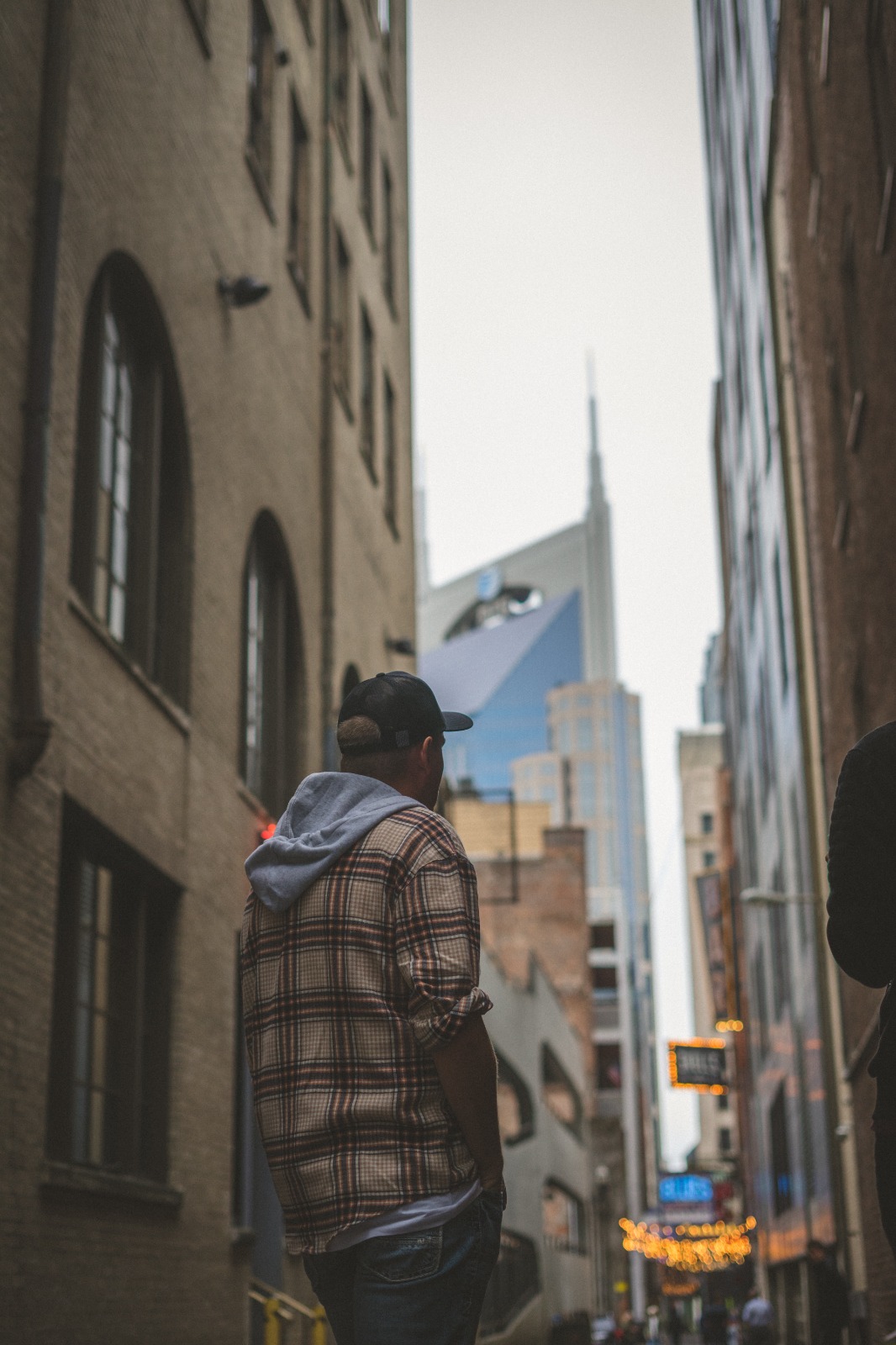 Person wearing a plaid jacket and cap walking in a city alley with tall buildings and a skyscraper in the background.