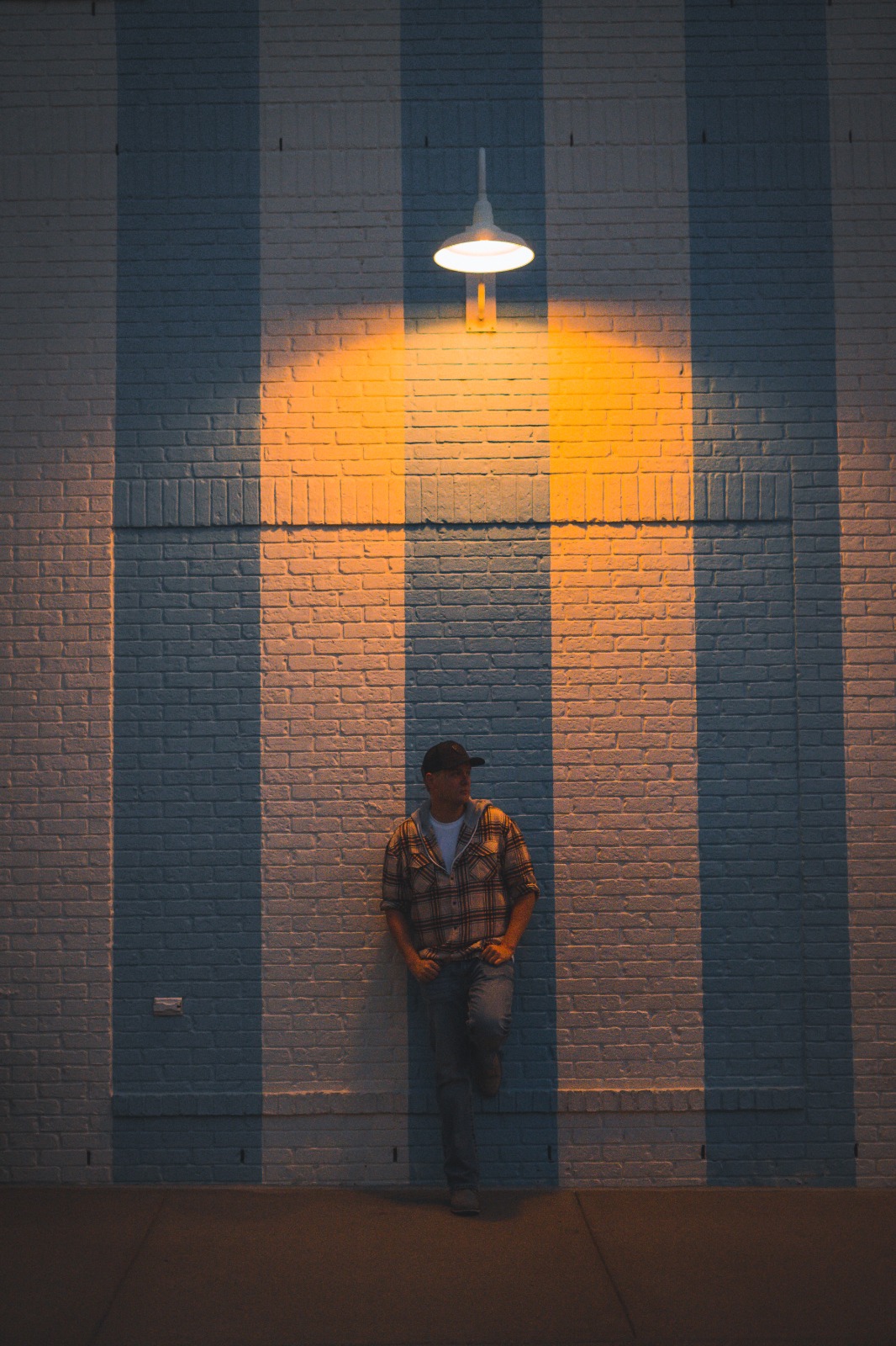 Person standing against a wall illuminated by a hanging light, with vertical blue and white stripes, in a dimly lit setting.