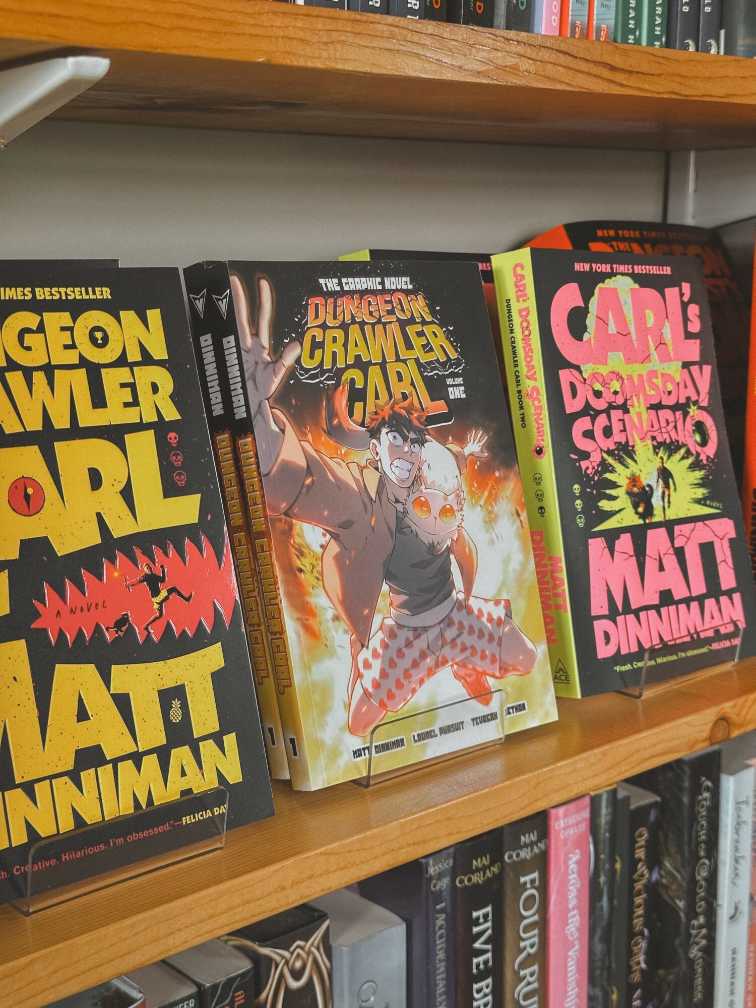 Three graphic novels on a bookshelf, featuring colorful covers with characters and titles, including 'Dungeon Crawler Carl' in the center.