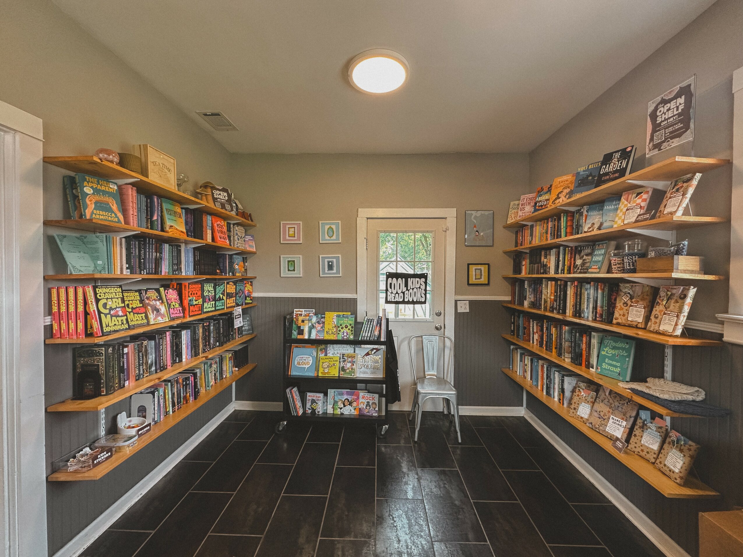 Bookshelves filled with books line both sides of a small room, with a window and door at the back, and a chair in front.