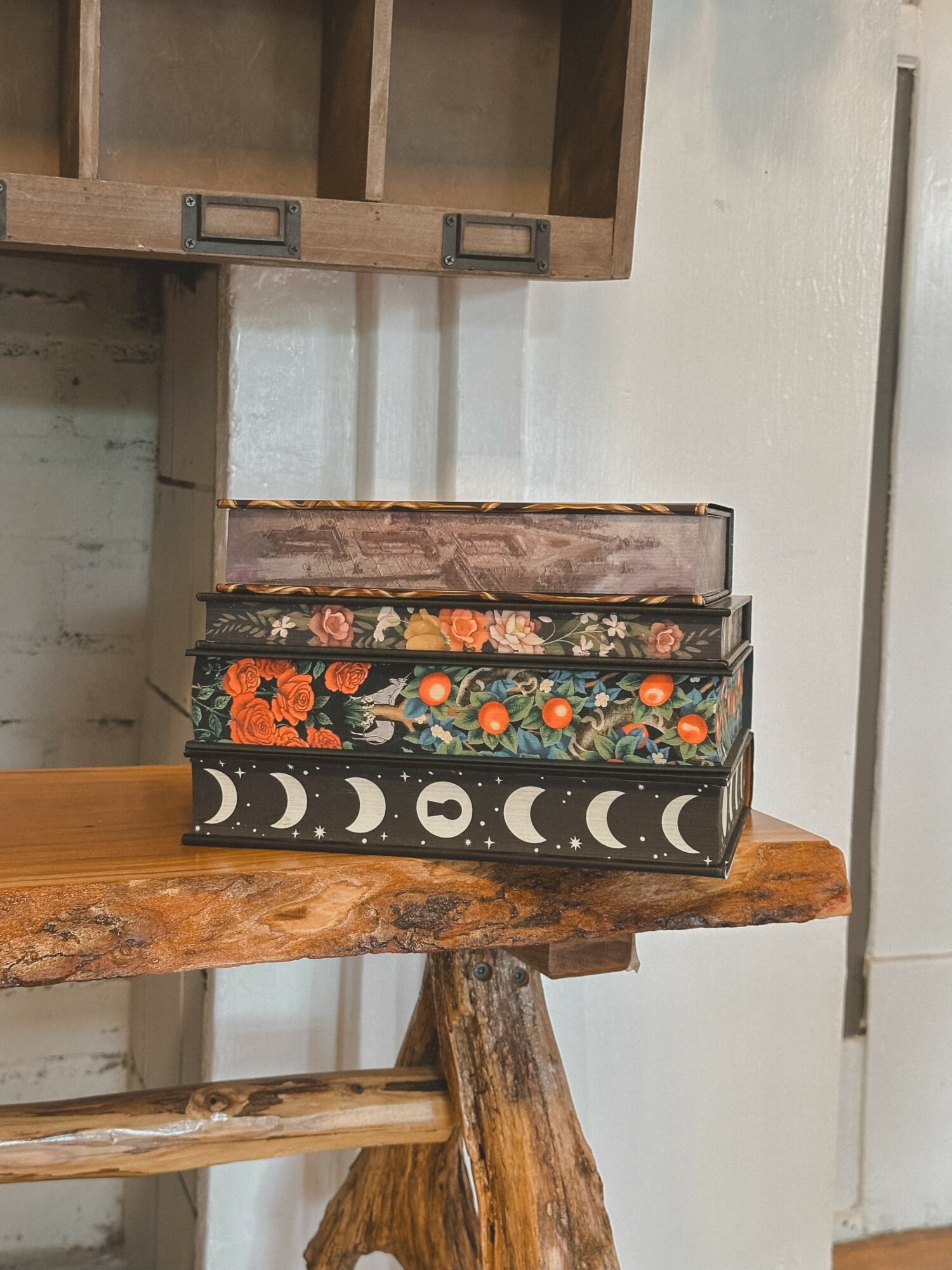 Three decorative boxes stacked on a wooden table, with a wall and shelf in the background.