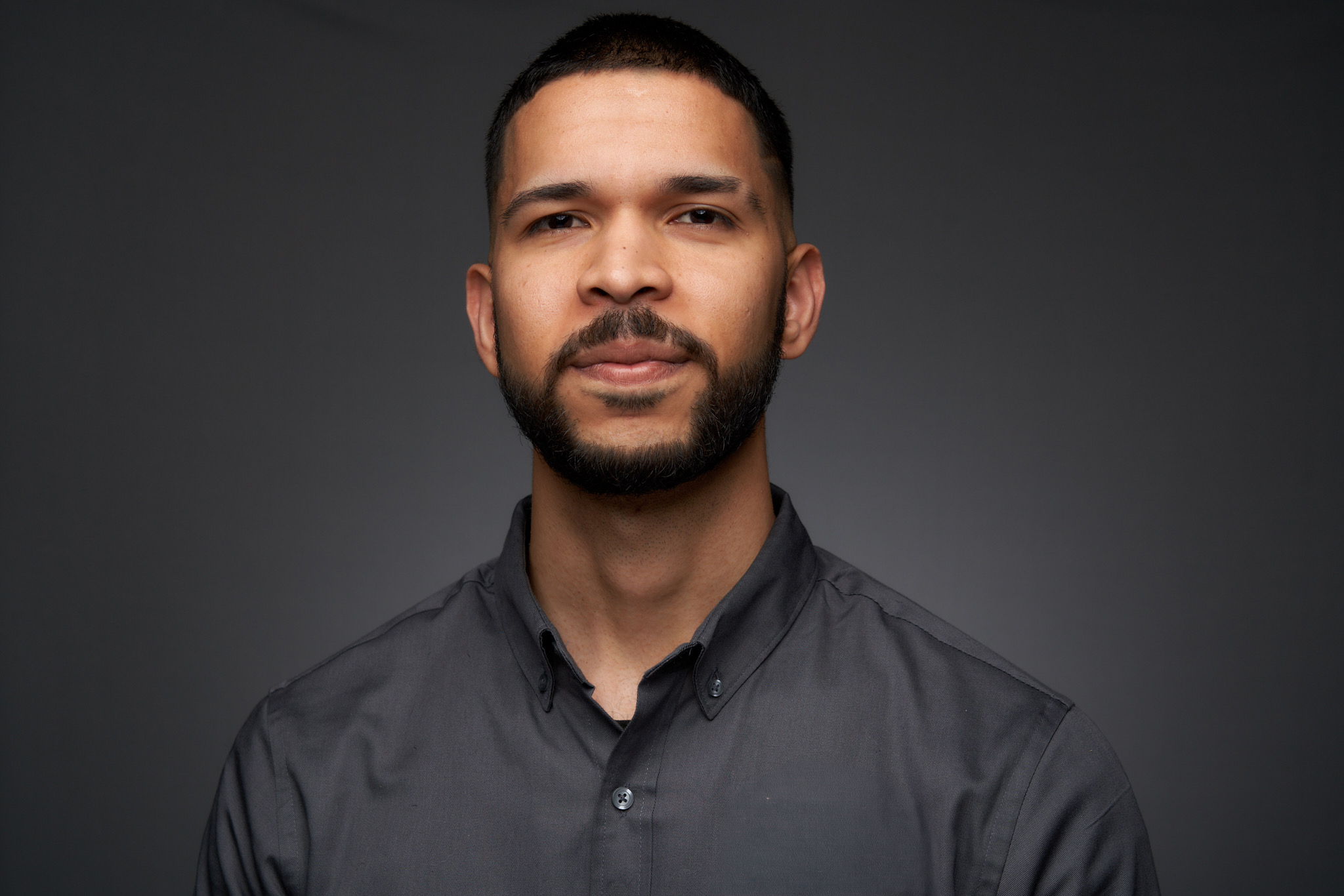 Portrait of a man with short dark hair and a beard, wearing a dark shirt, against a dark background.