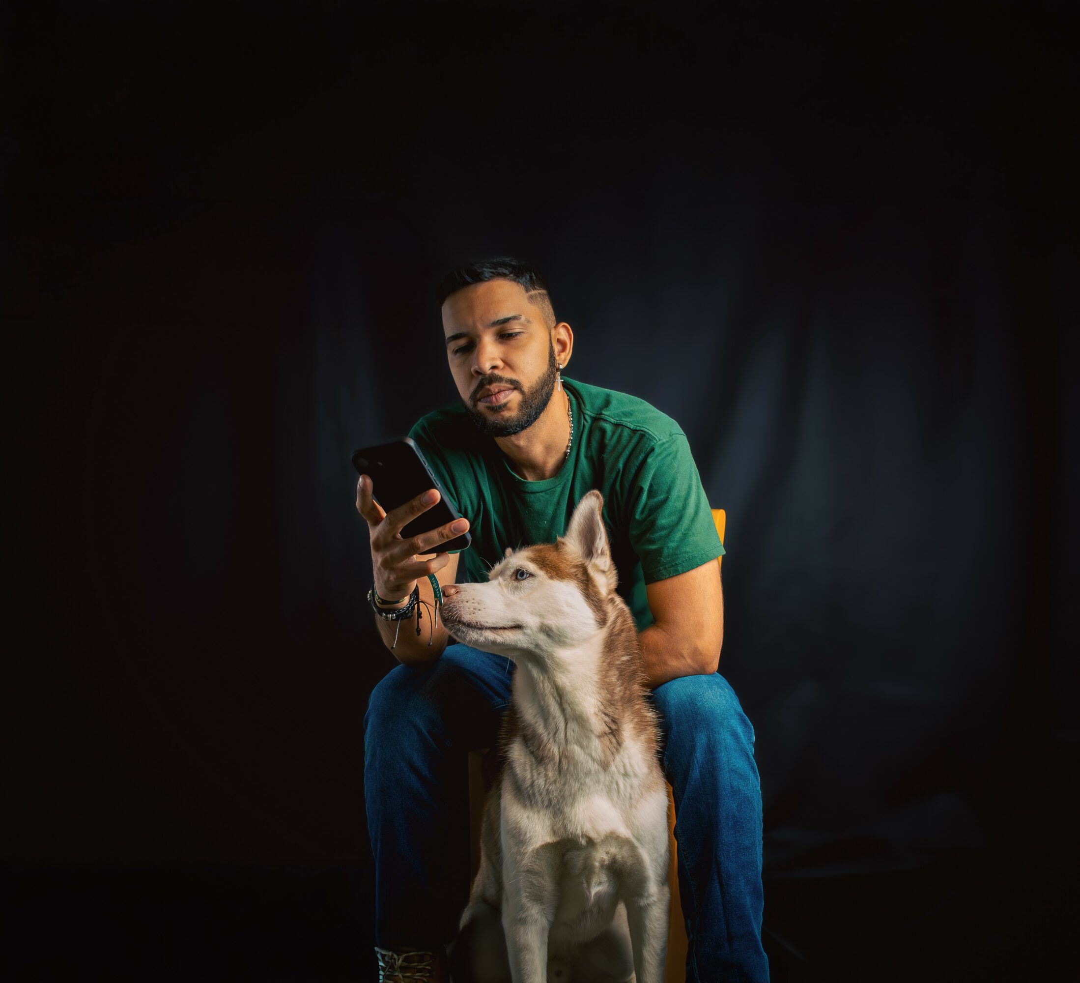 Man sitting with a dog, looking at a phone, against a dark background.