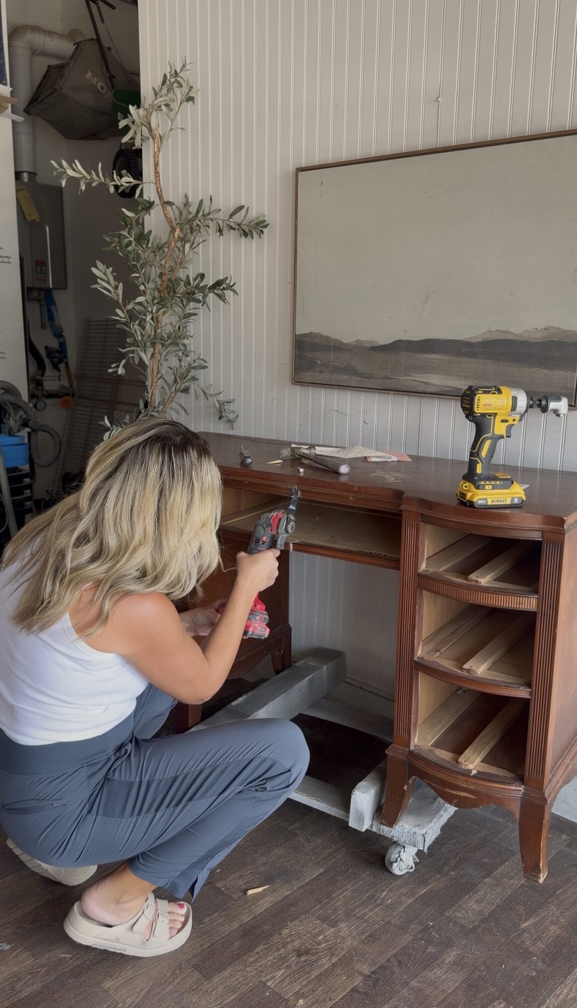 Woman crouching and taking a photo of a wooden cabinet with a yellow drill on top, in a room with a mirror and plant.