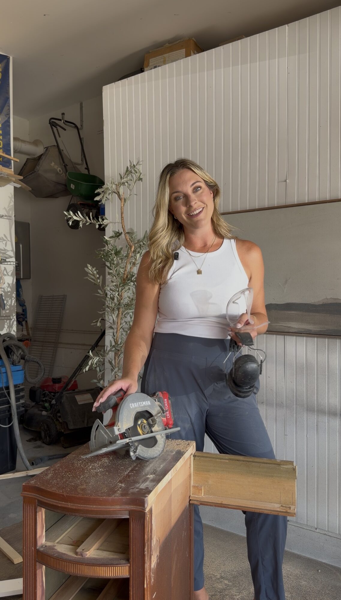 Woman with long blonde hair smiling, holding a camera, standing in a garage with a table saw and storage cabinets.
