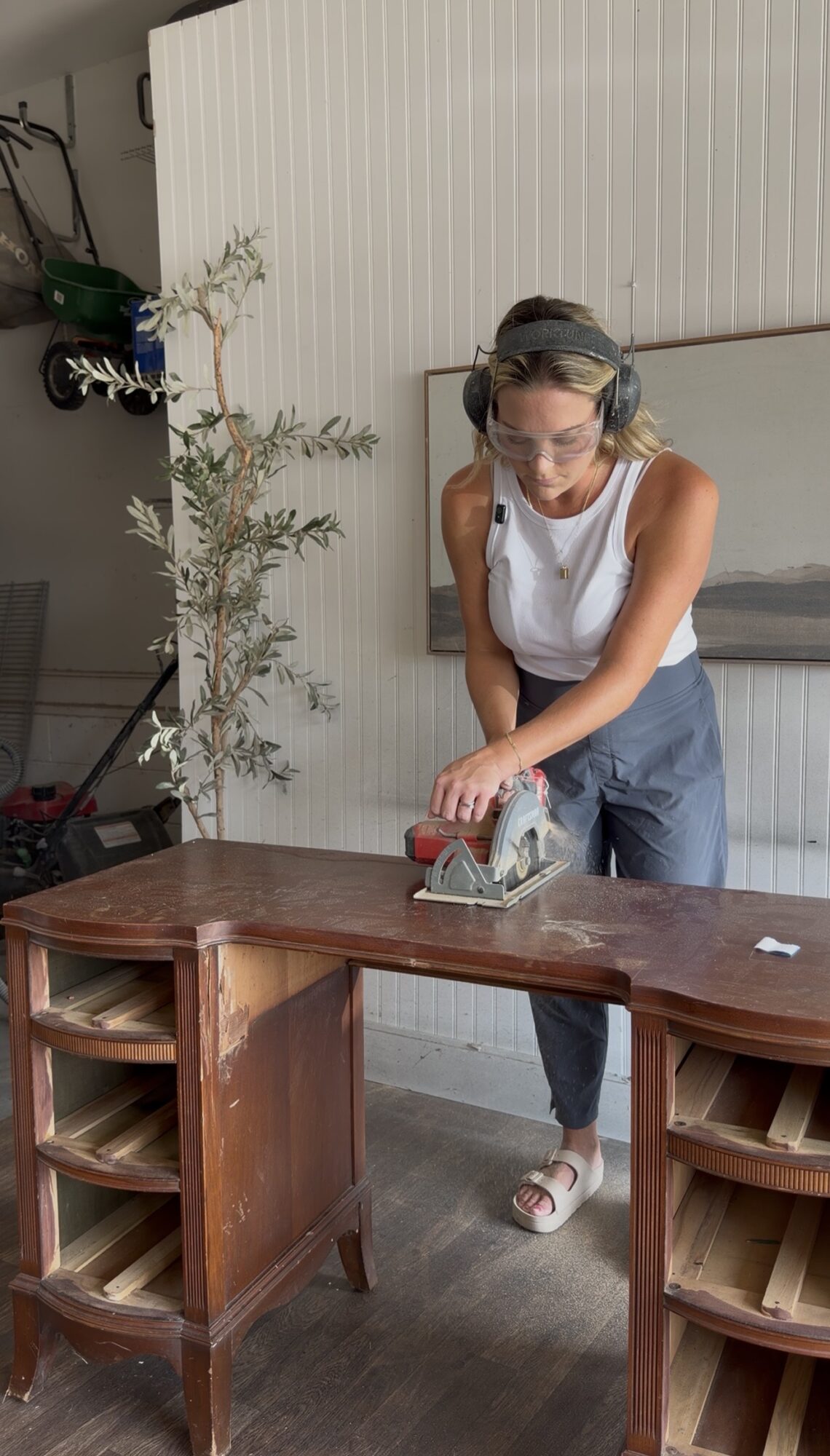 Woman using a handheld power tool on a wooden table, wearing safety goggles and headphones, in a room with a white wall and plant.