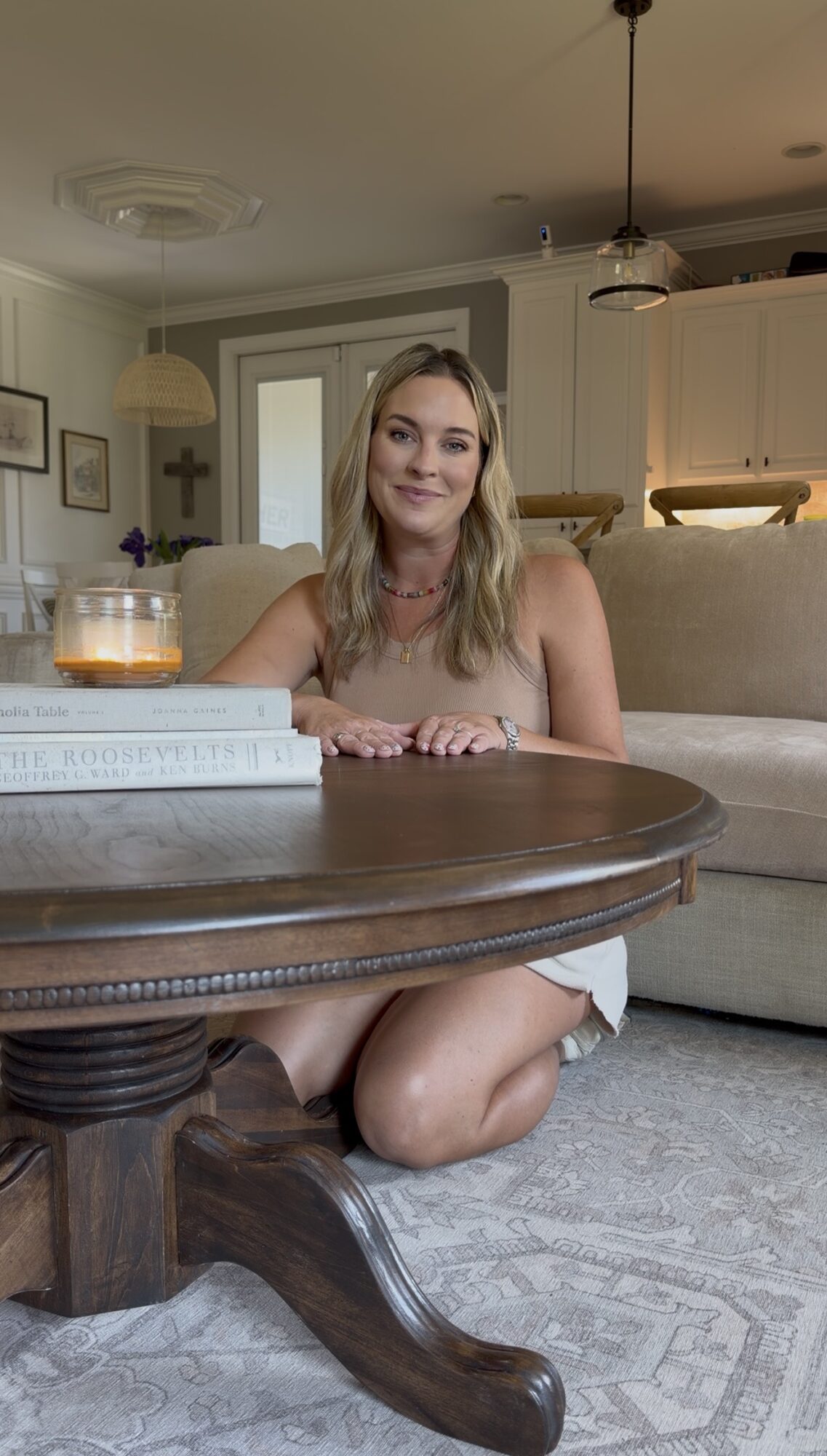Young woman with blonde hair smiling, sitting on her knees at a round wooden table in a living room, with a drink and books.