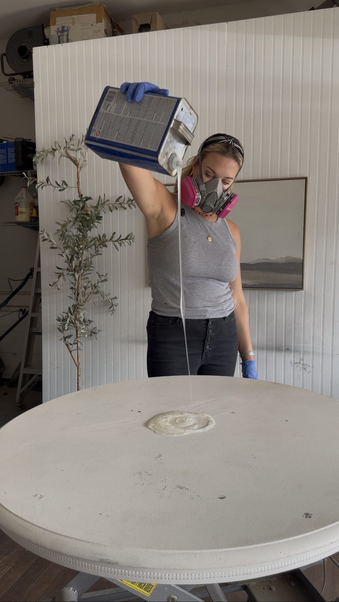 Person wearing a mask and gloves pouring liquid from a container into a large round object, with a white wall and plant in background.