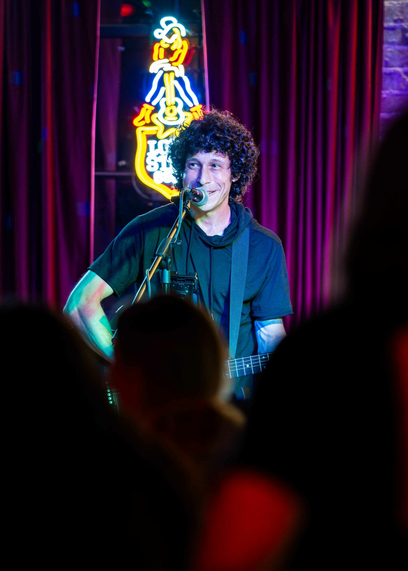 Musician with curly hair performs on stage with a guitar, microphone, and a neon sign in the background, audience in foreground.