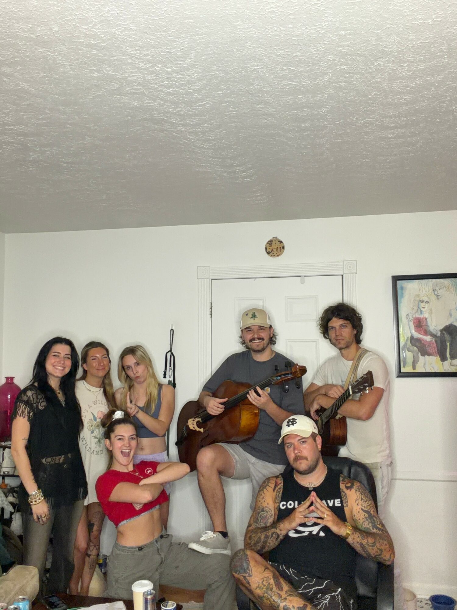 Group of seven people in a room, some holding guitars, with a door and framed picture on the wall behind them.