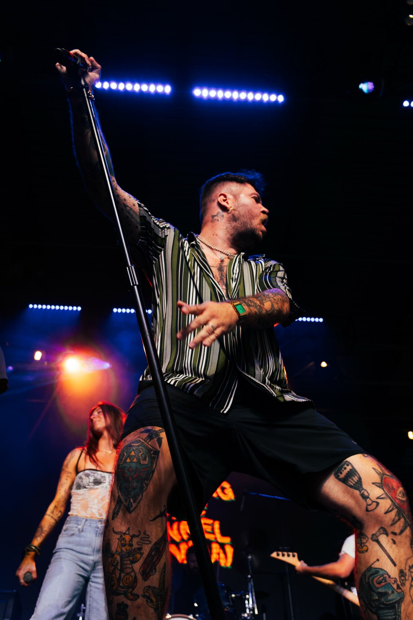 Male performer singing on stage with a microphone, wearing a striped shirt and shorts, tattoos visible on legs, stage lights in background.