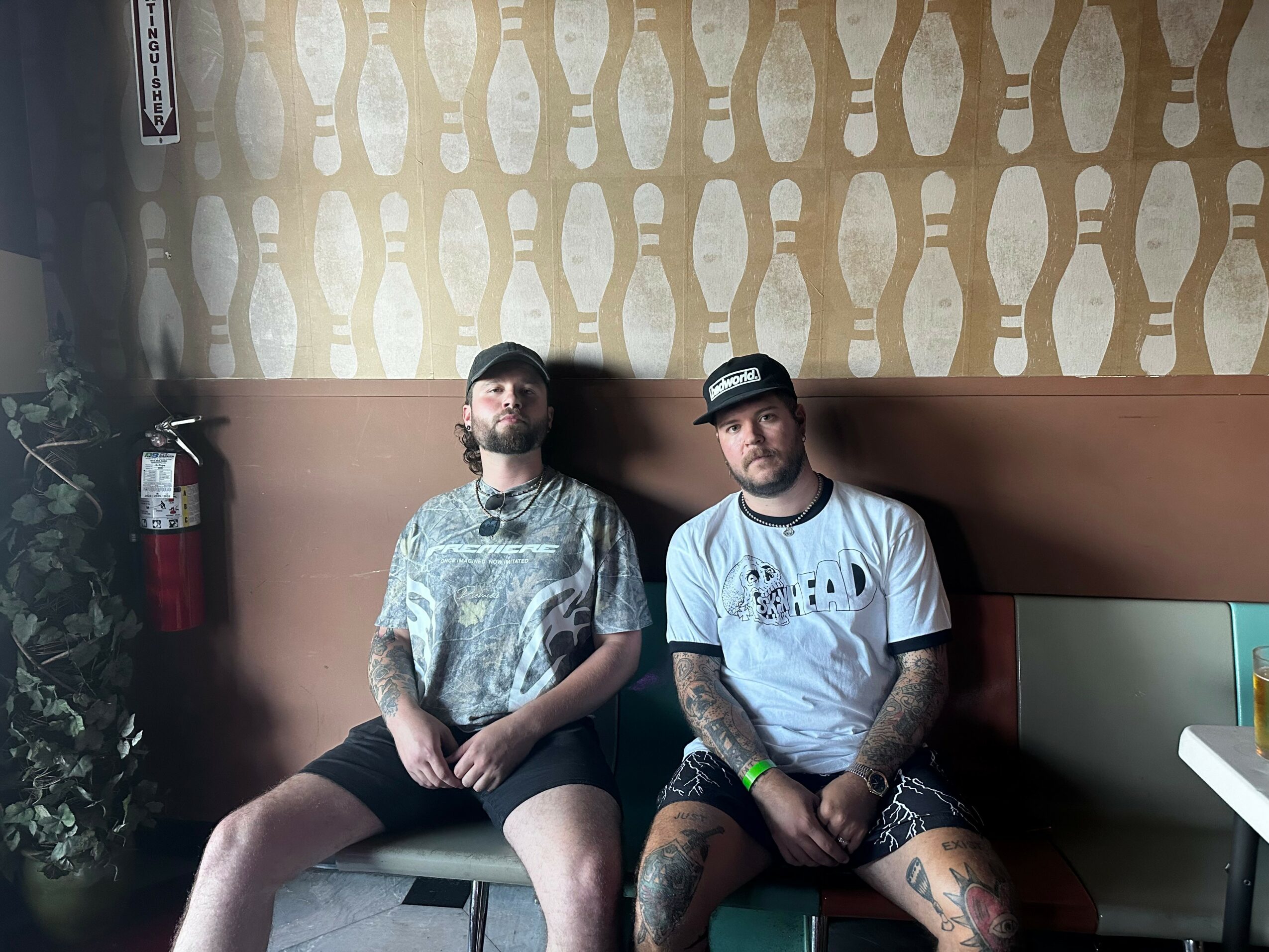 Two men sitting on a bench indoors, one with a beard and tattoos, wearing a patterned shirt and shorts, the other with a cap and tattoos, wearing a t-shirt and shorts.