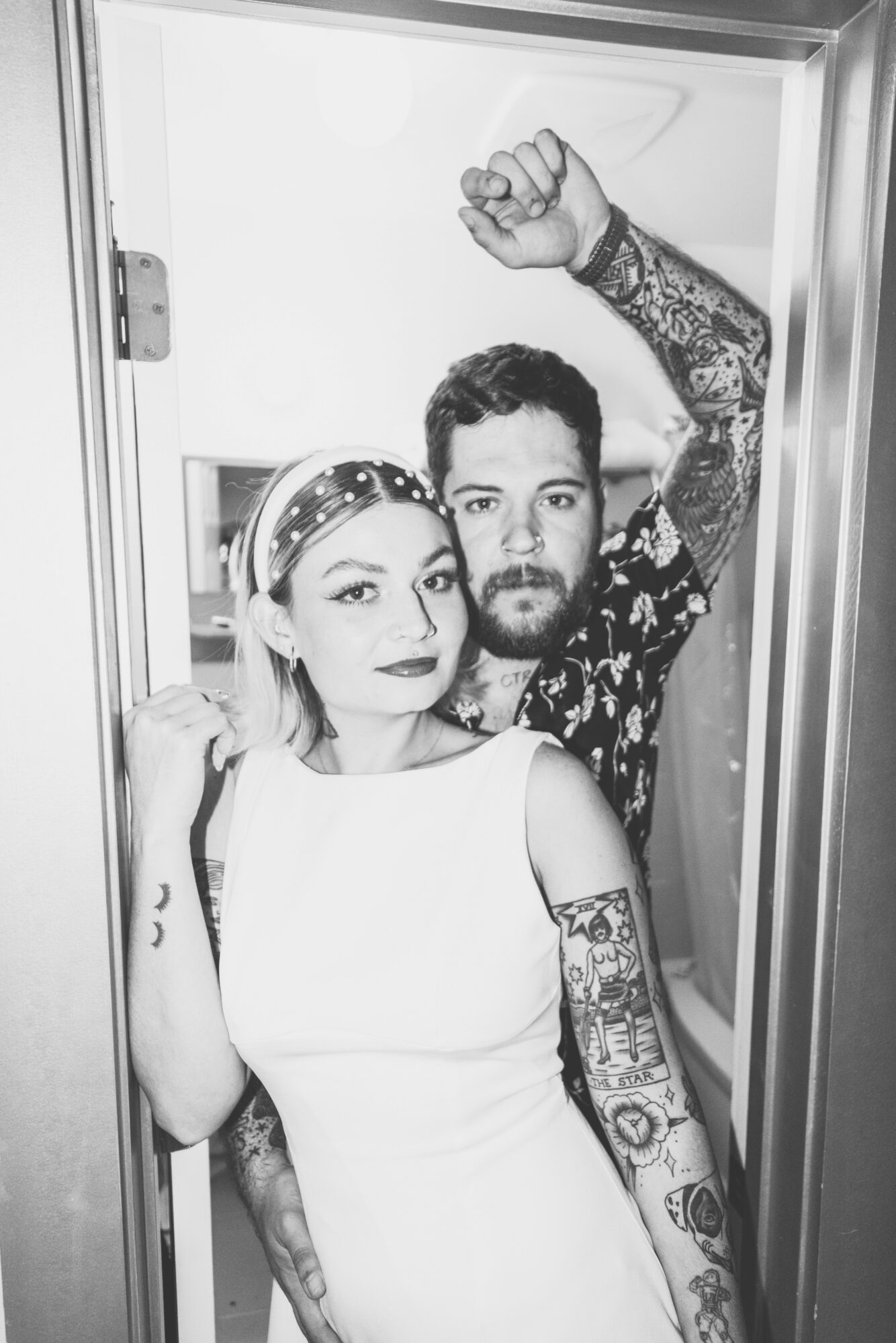Two people with tattoos pose in a small space, one with a raised fist, both looking at the camera.