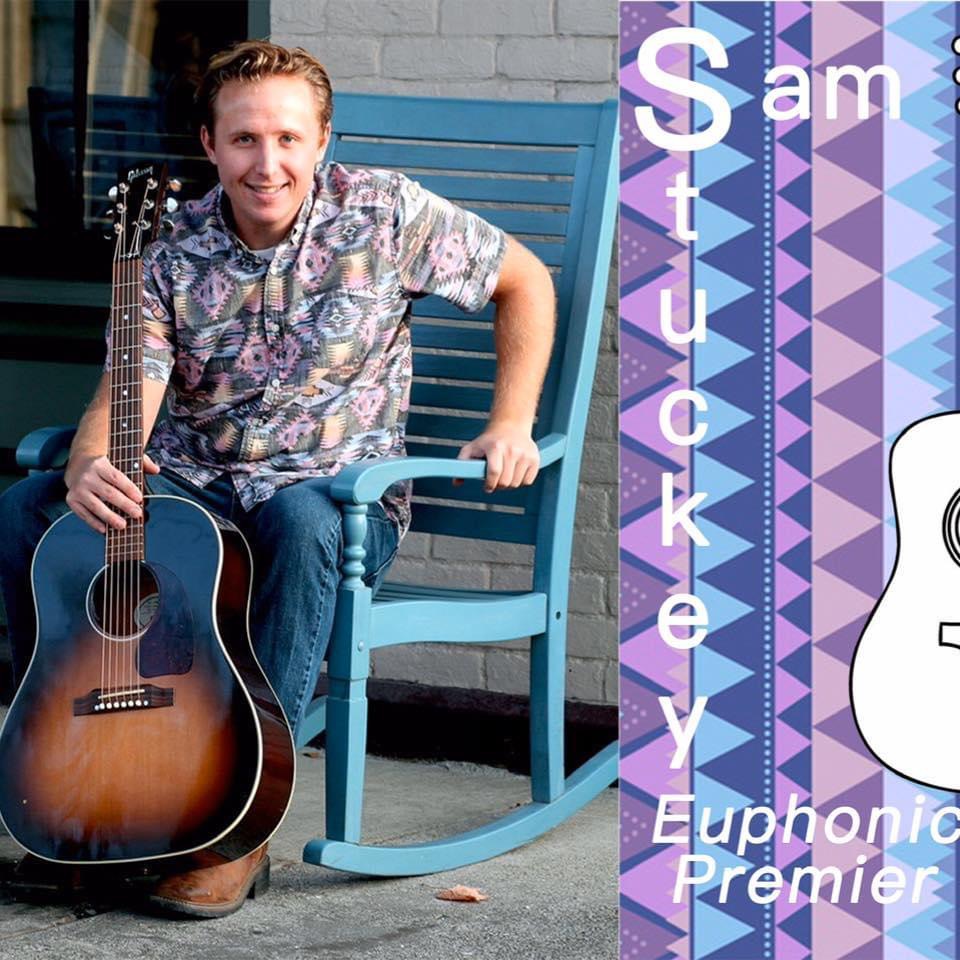 Person sitting on a blue rocking chair holding a guitar, smiling outdoors next to a colorful sign that reads 'Sam Stuckey, Euphonium Premier'.