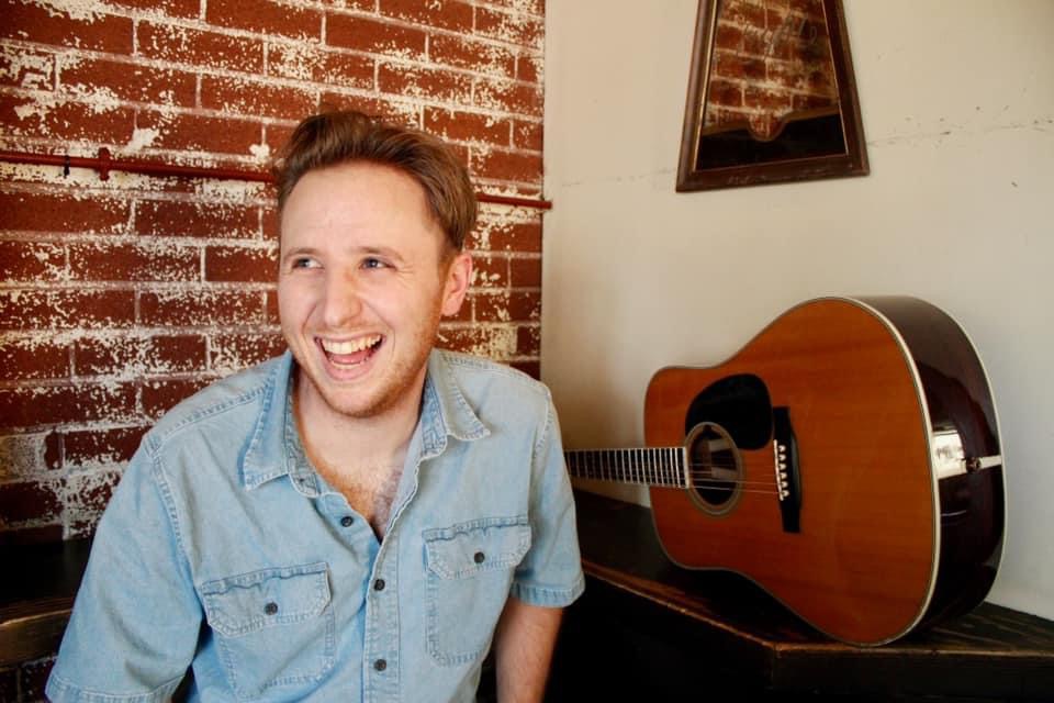 Smiling man with short hair and a light blue shirt sitting indoors next to an acoustic guitar on a table, brick wall background.