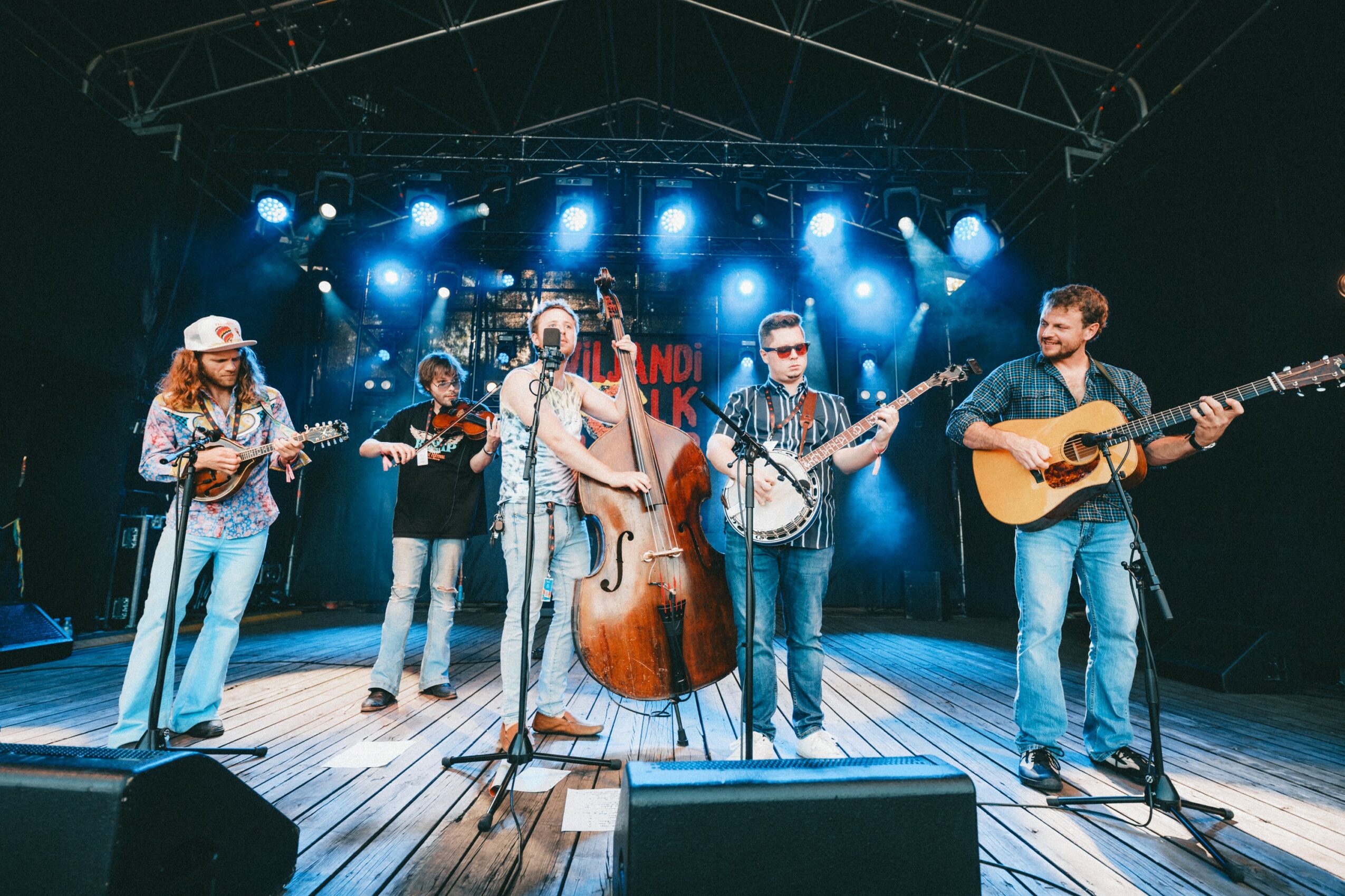 Six musicians perform on stage with blue lighting, playing instruments including guitar, bass, and violin, in a concert setting.