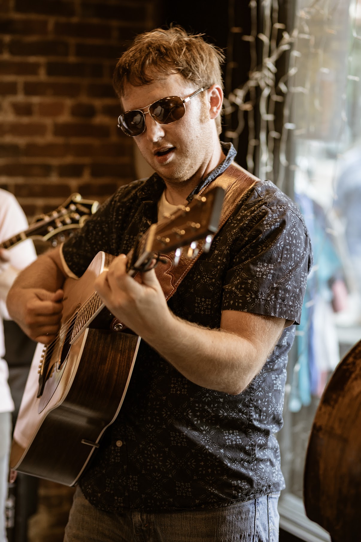 Young man playing guitar, wearing sunglasses, in a dimly lit indoor setting with a brick wall background.