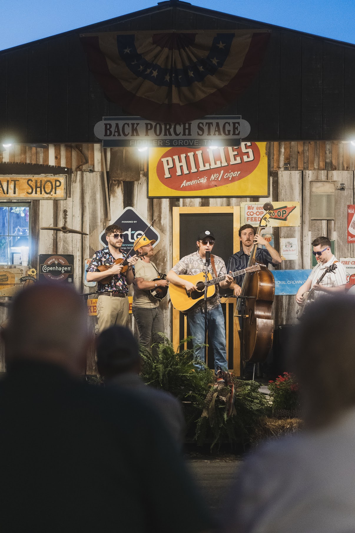 Band performs on stage with audience in foreground, wooden building backdrop, signs, and banners, outdoors during daytime.