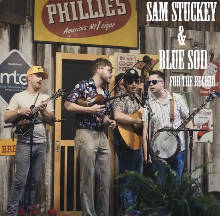 Four musicians perform outdoors with guitars, wearing casual clothing and sunglasses, in front of a wooden building with signs.