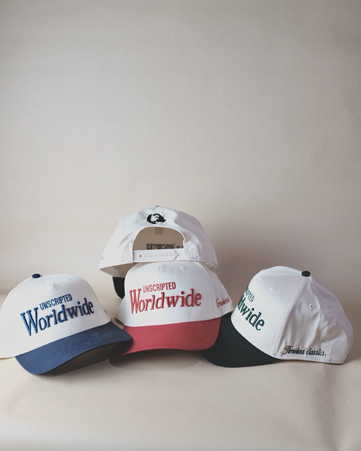 Three baseball caps with text, one with a white front and blue brim, one with a white front and red brim, and one with a white front and black brim, all with adjustable straps. The caps have text related to being worldwide.