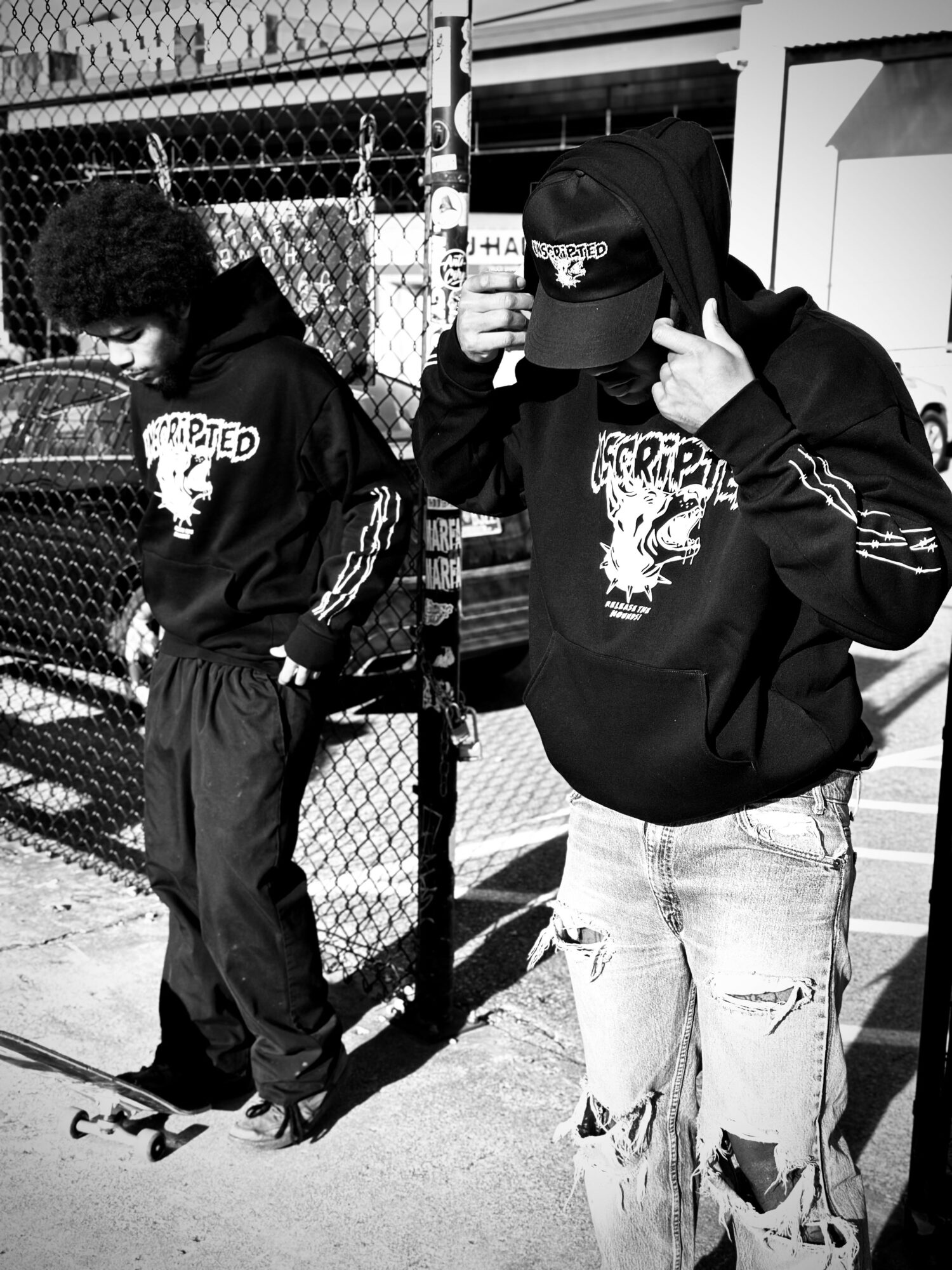Two young people standing outdoors near a chain-link fence, one holding a phone, both wearing hoodies with graphic designs.