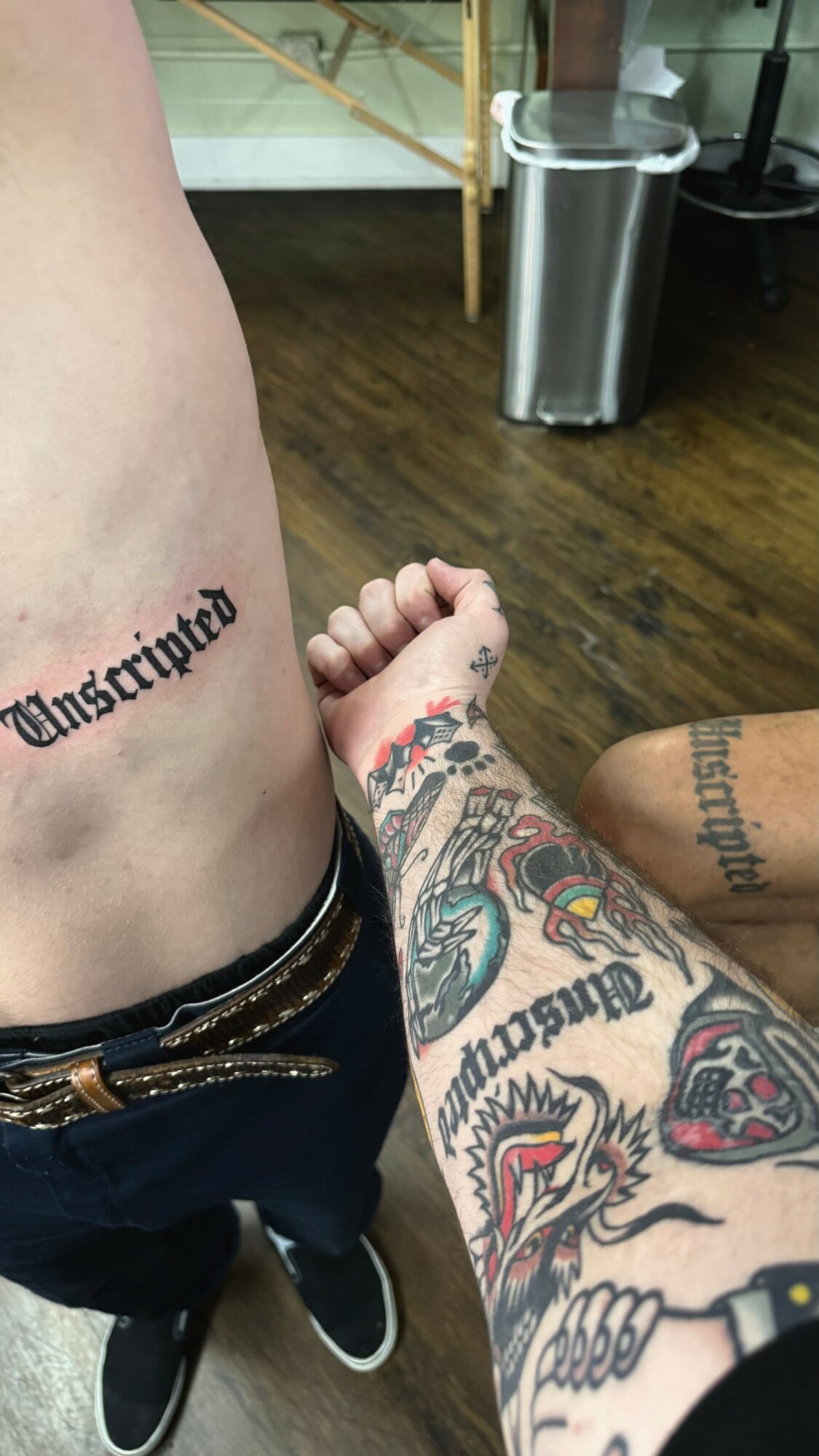 Close-up of a person's side and arm with tattoos, showing a tattooed arm reaching towards the side, background includes a trash can and furniture.