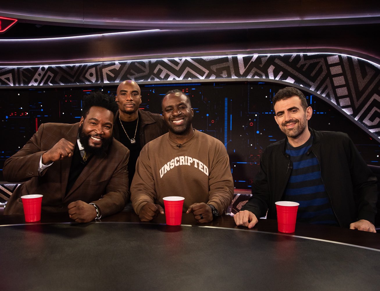 Four men sitting at a table with red cups, smiling, in a studio with geometric background design.
