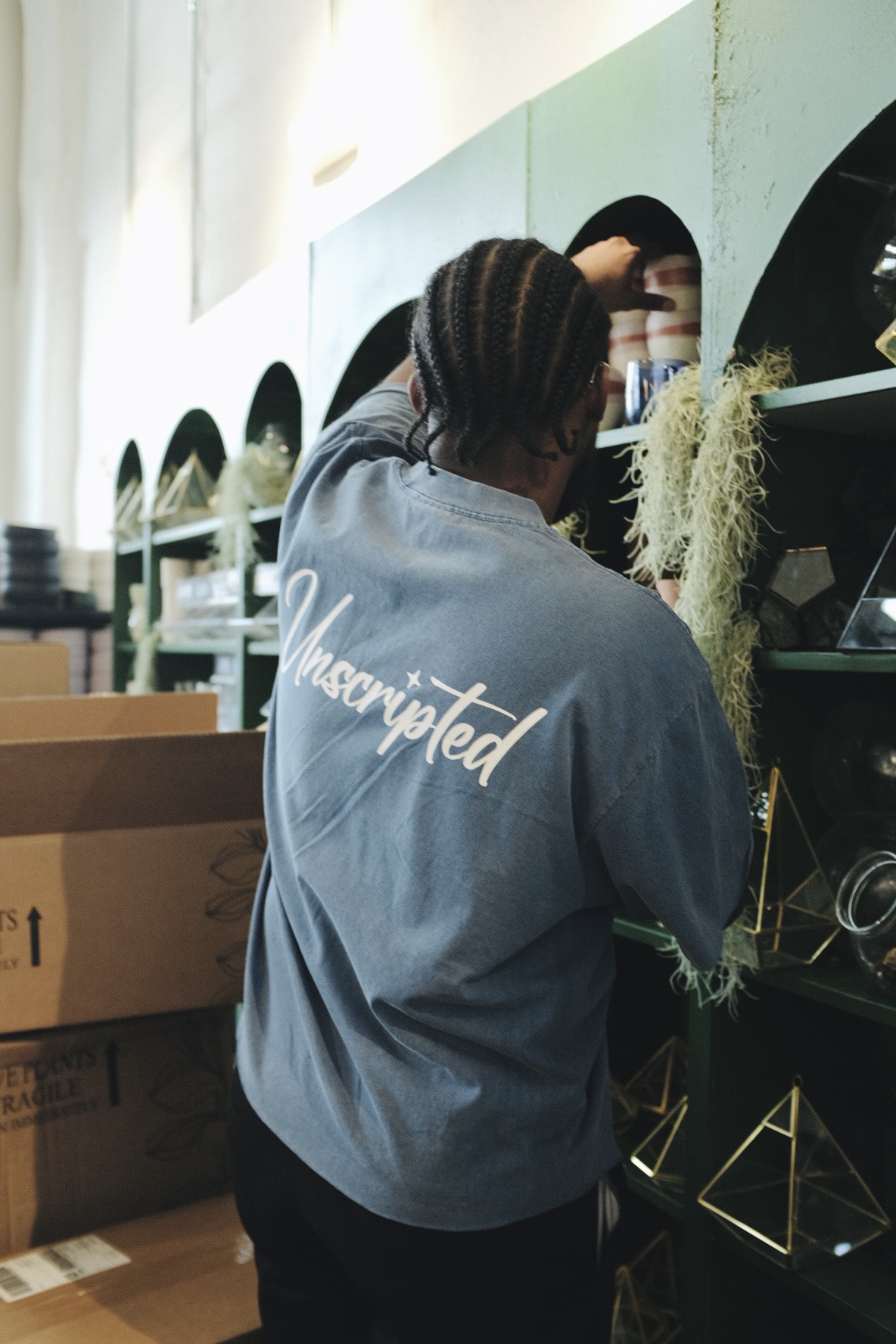 Person with braided hair wearing a gray shirt with white text, facing shelves in a store, with boxes nearby.