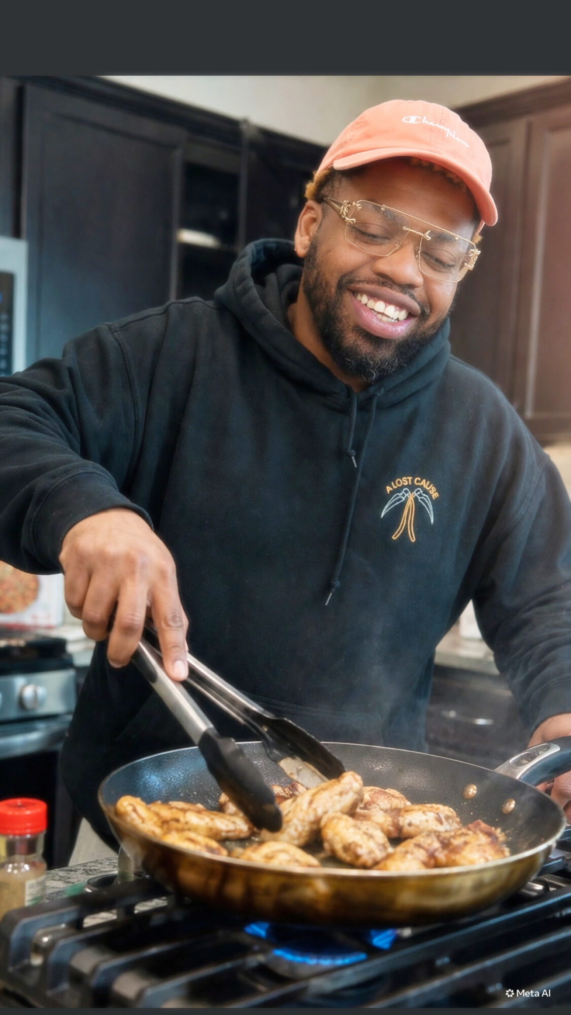 Man cooking chicken in a frying pan, smiling, wearing glasses, a pink cap, and a black hoodie in a kitchen.